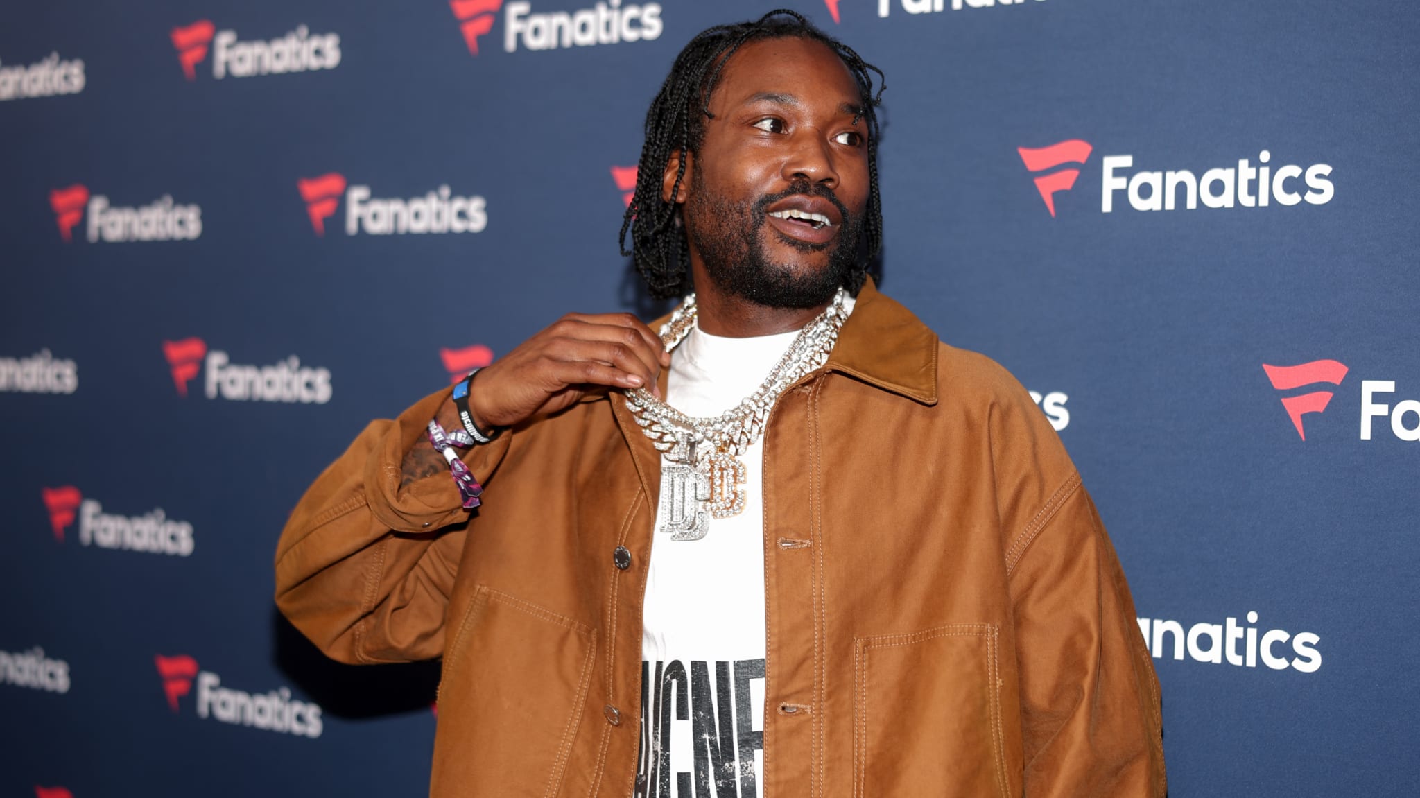 A man in a brown jacket and white shirt, wearing a large chain necklace, stands in front of a Fanatics-branded backdrop.