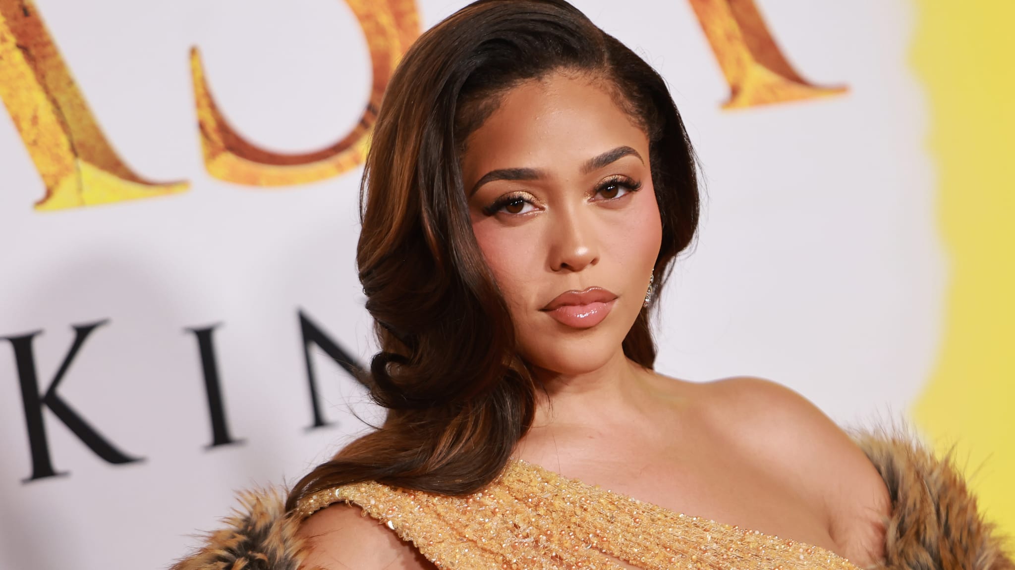 Jordyn Woods in a glamorous gold dress with a fur coat poses on a red carpet with a yellow and white background.