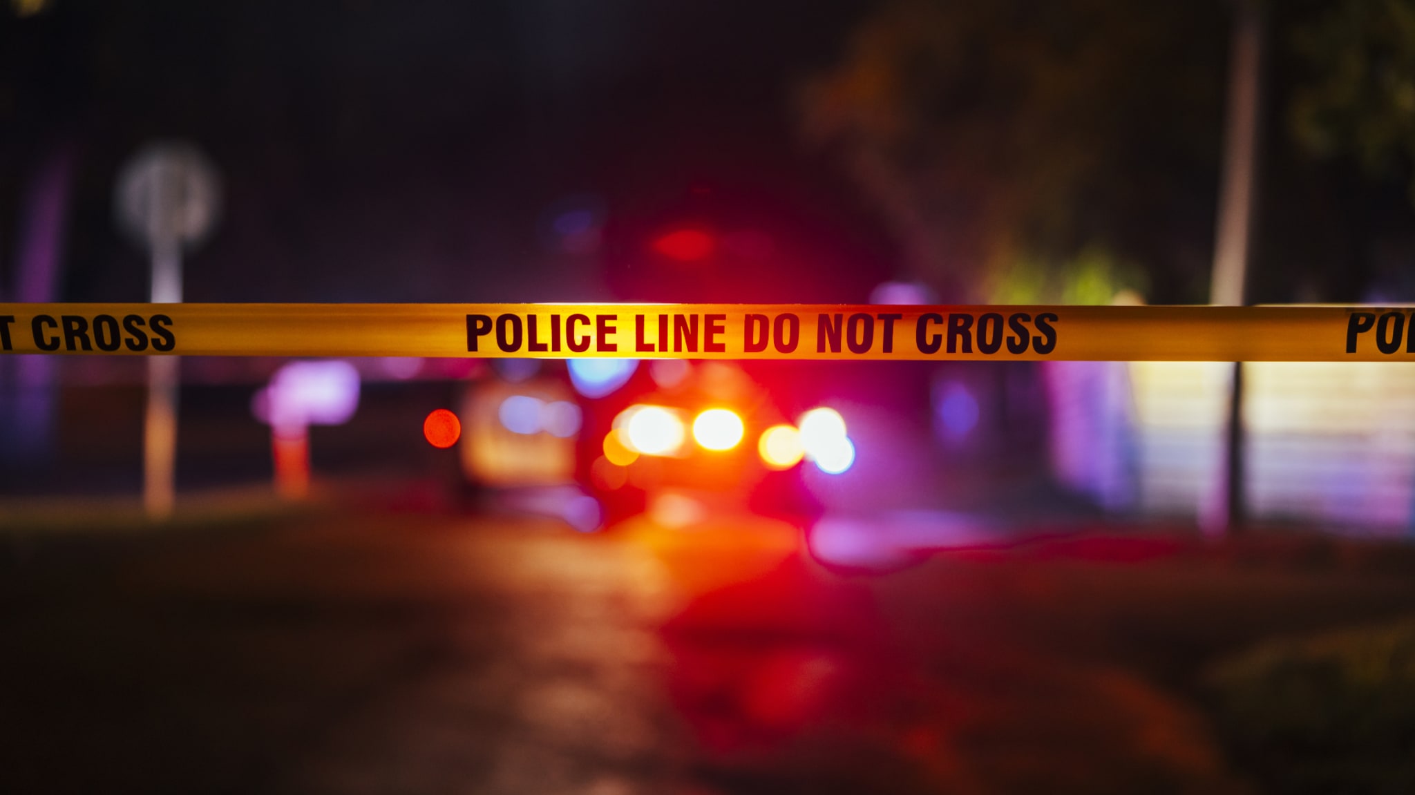 Police tape with "POLICE LINE DO NOT CROSS" text, blurred background with flashing lights, indicating a crime scene or emergency situation.