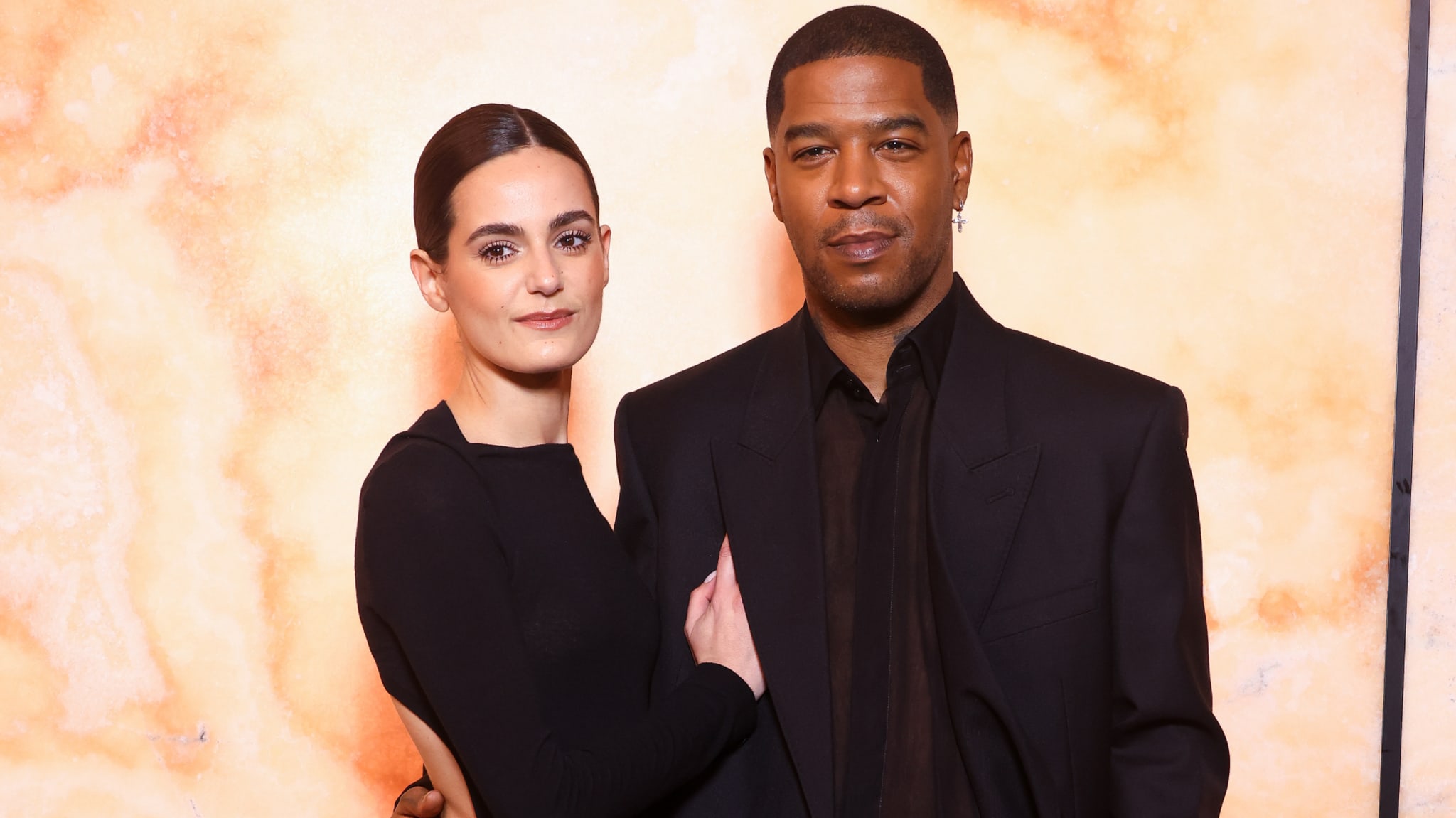 A woman in a black dress and a man in a black suit pose together against a marbled background.