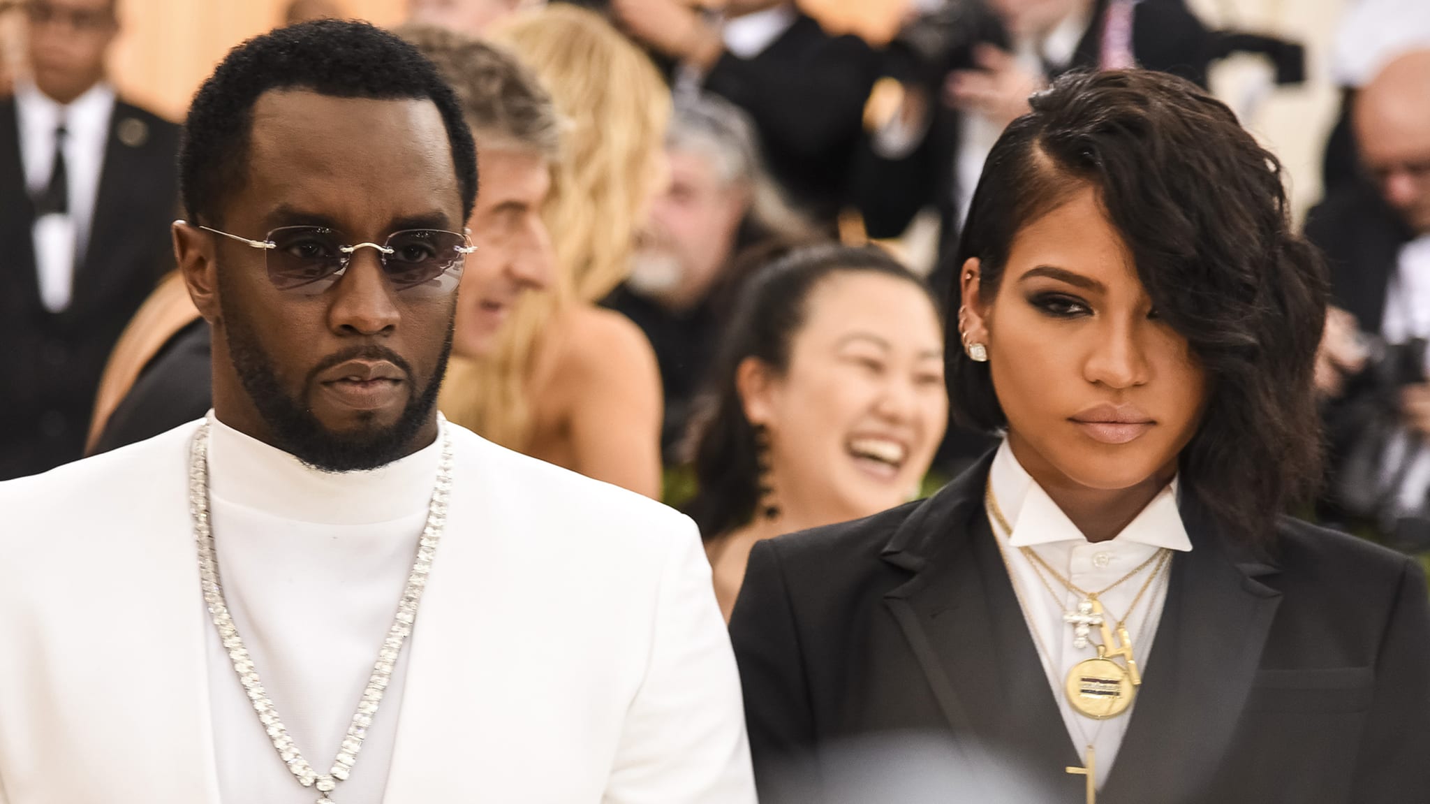 Sean "Diddy" Combs and Cassie at a formal event, with Diddy in a white suit and sunglasses, and Cassie in a black suit.