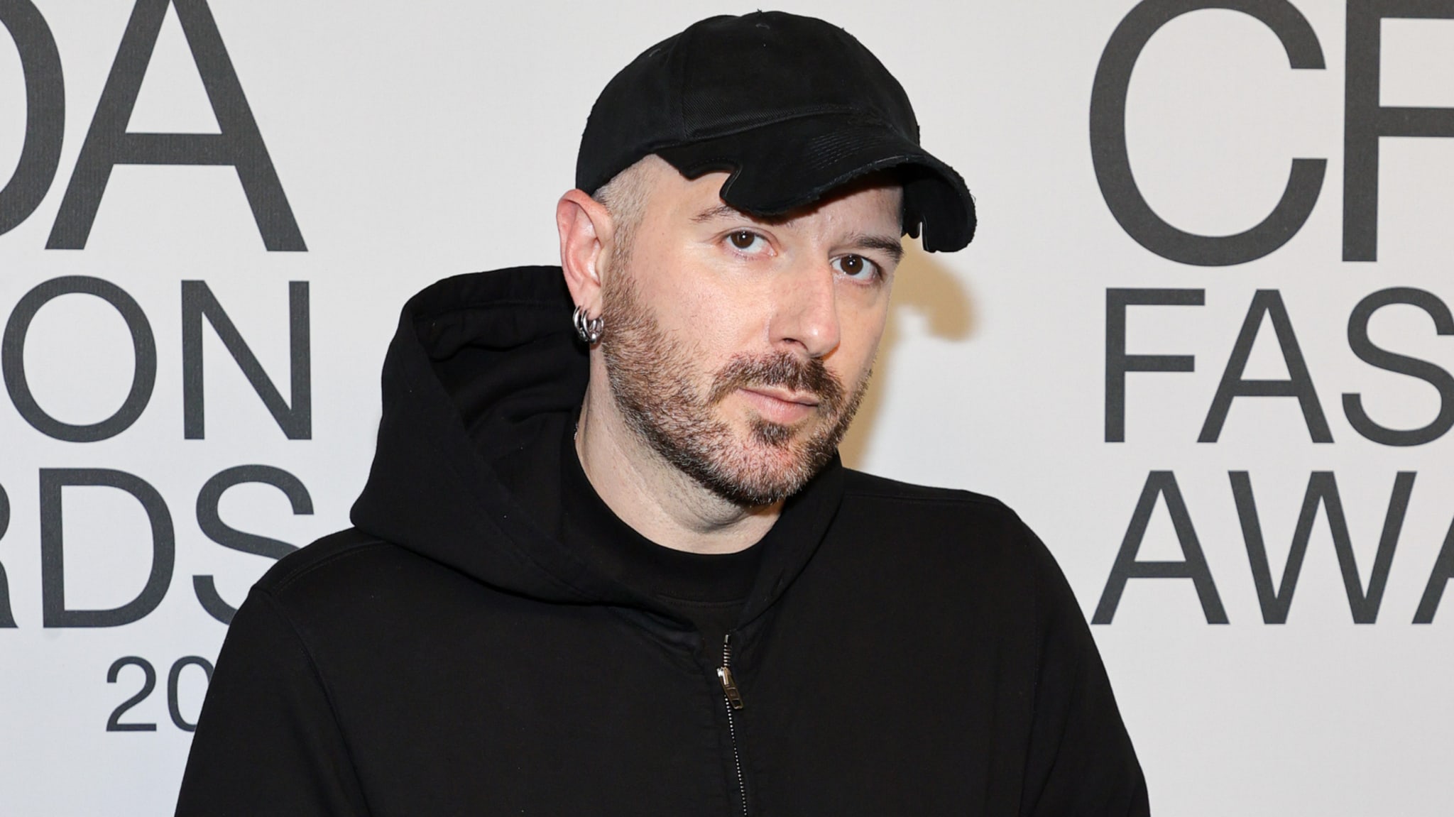 A man wearing a black hoodie and cap stands in front of a "CFDA Fashion Awards" backdrop.