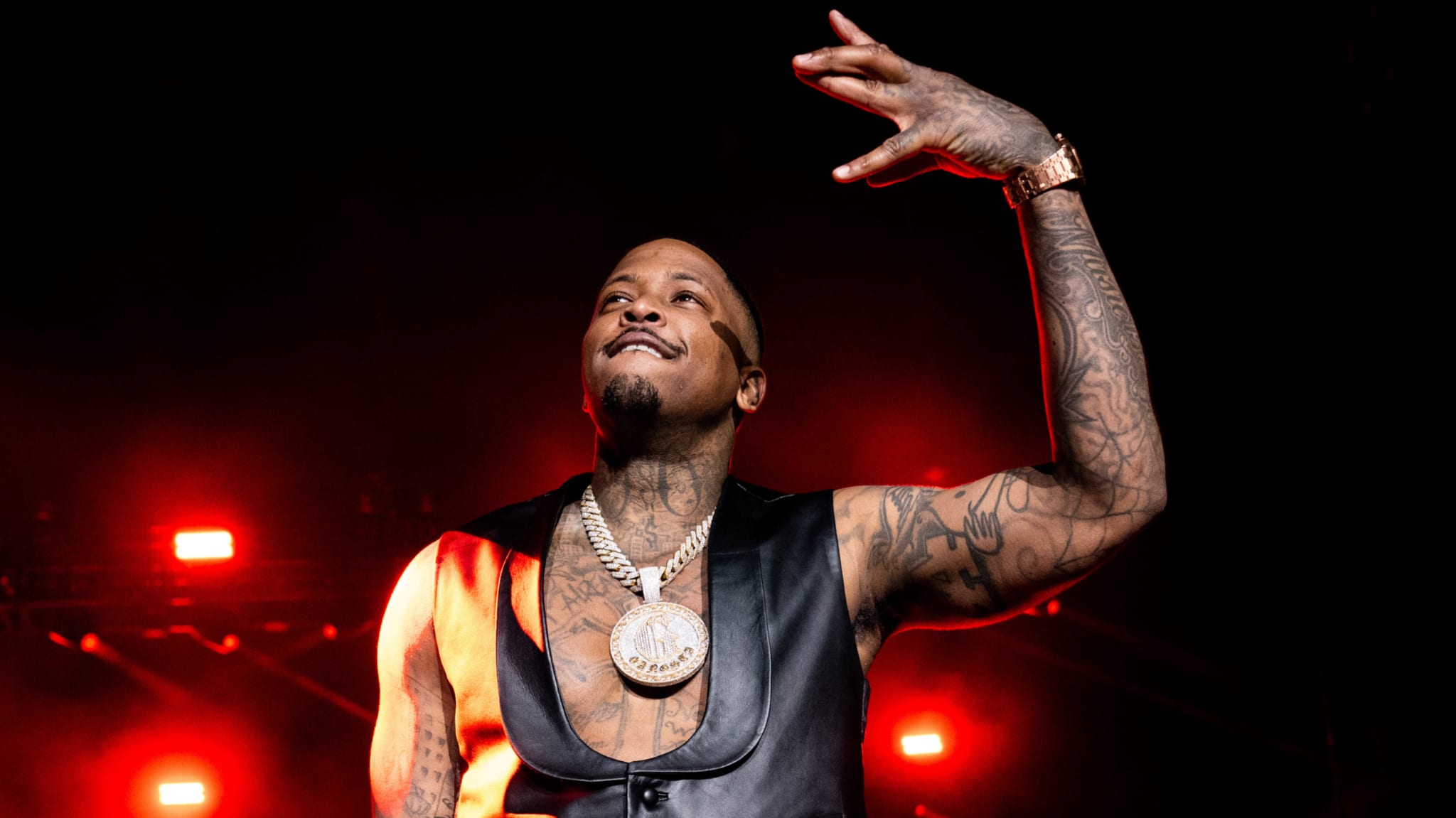 Rapper YG performing on stage, wearing a black vest and large chain, with red stage lights in the background.