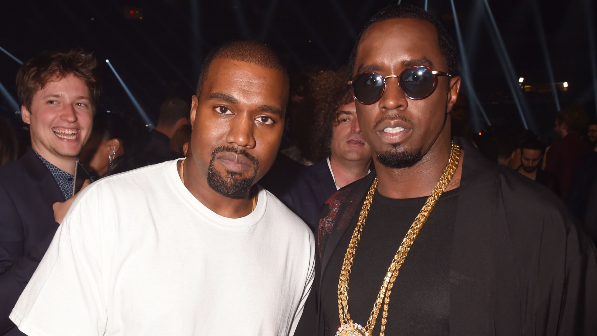 Kanye West in a white shirt and sunglasses-wearing Diddy with a gold chain, at an event with people in the background.