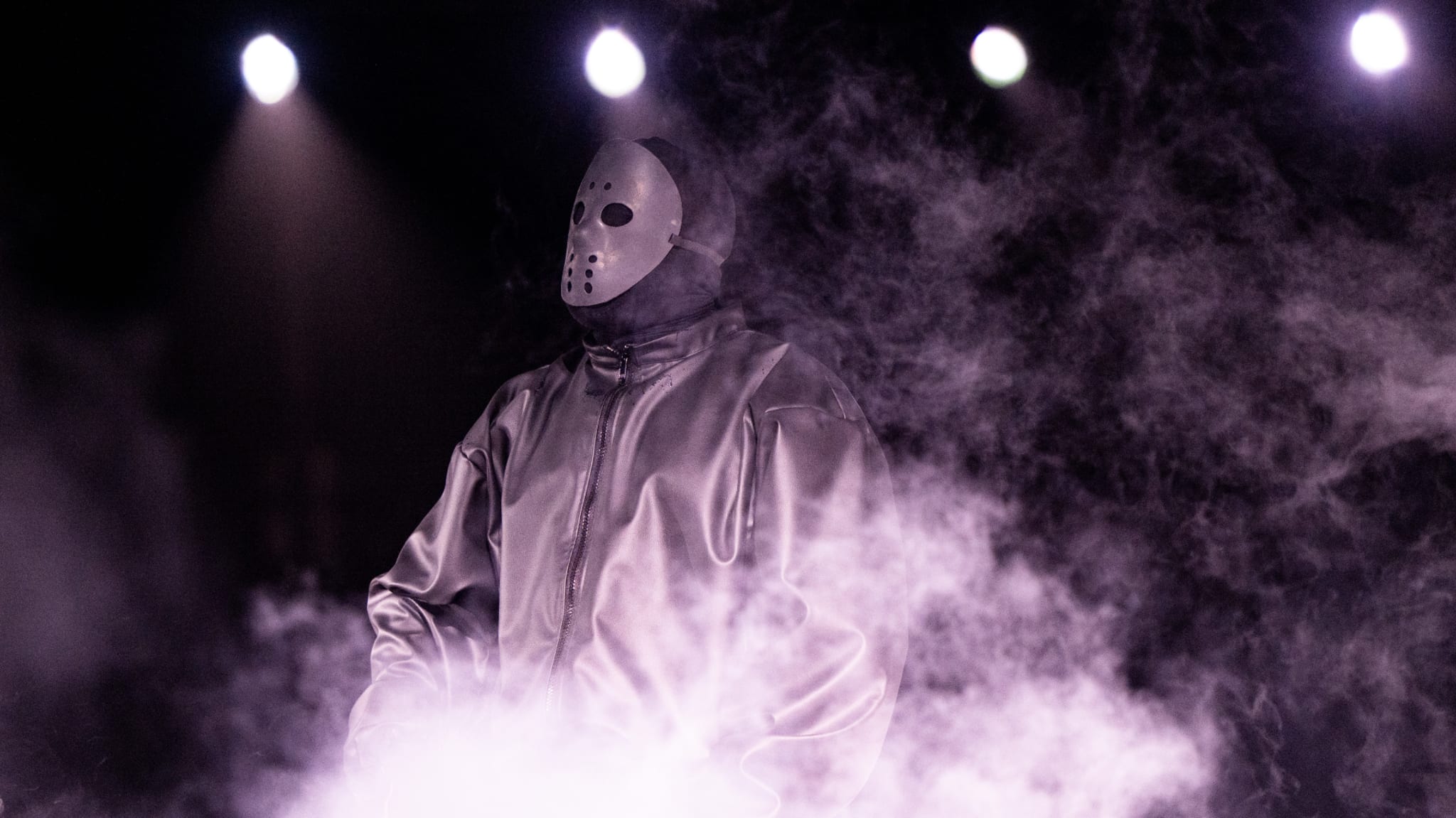 Ye wearing a hockey mask and a shiny jacket stands amid fog, with bright lights in the background.