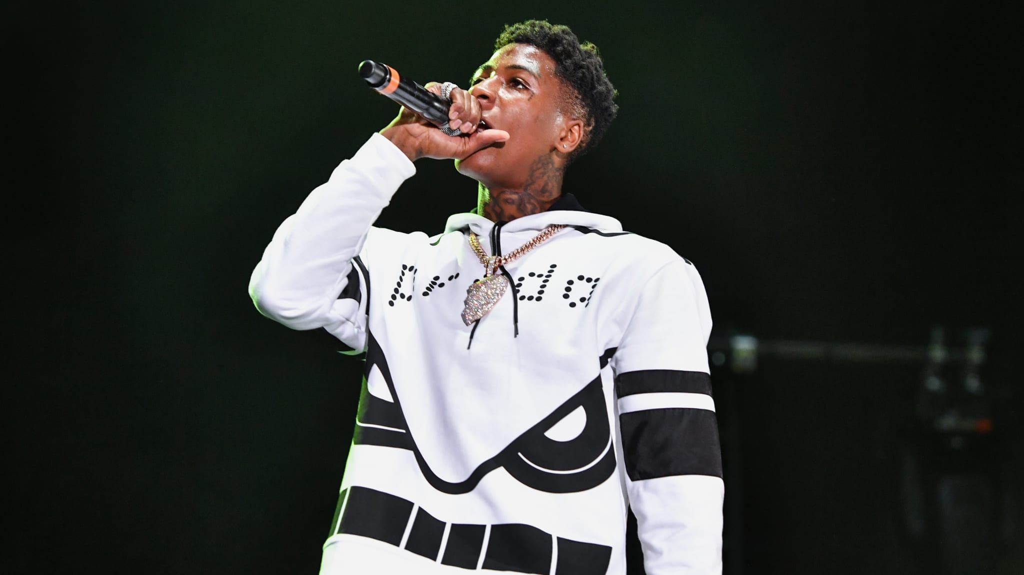 NBA YoungBoy performing on stage, wearing a white hoodie with black designs and holding a microphone.