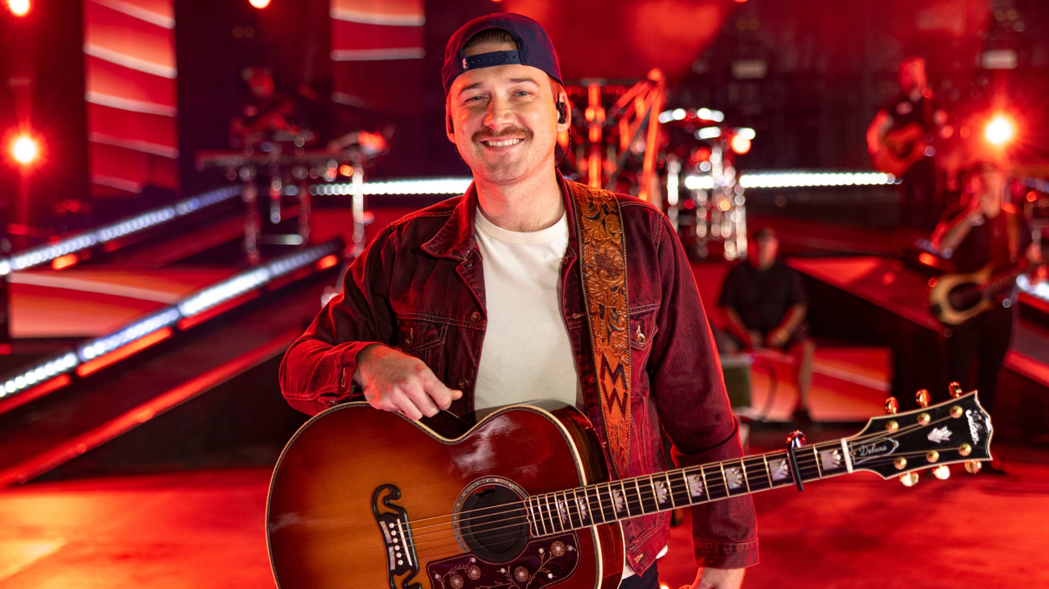 A man with a guitar on stage, wearing a cap and jacket, with red lighting and band equipment in the background.