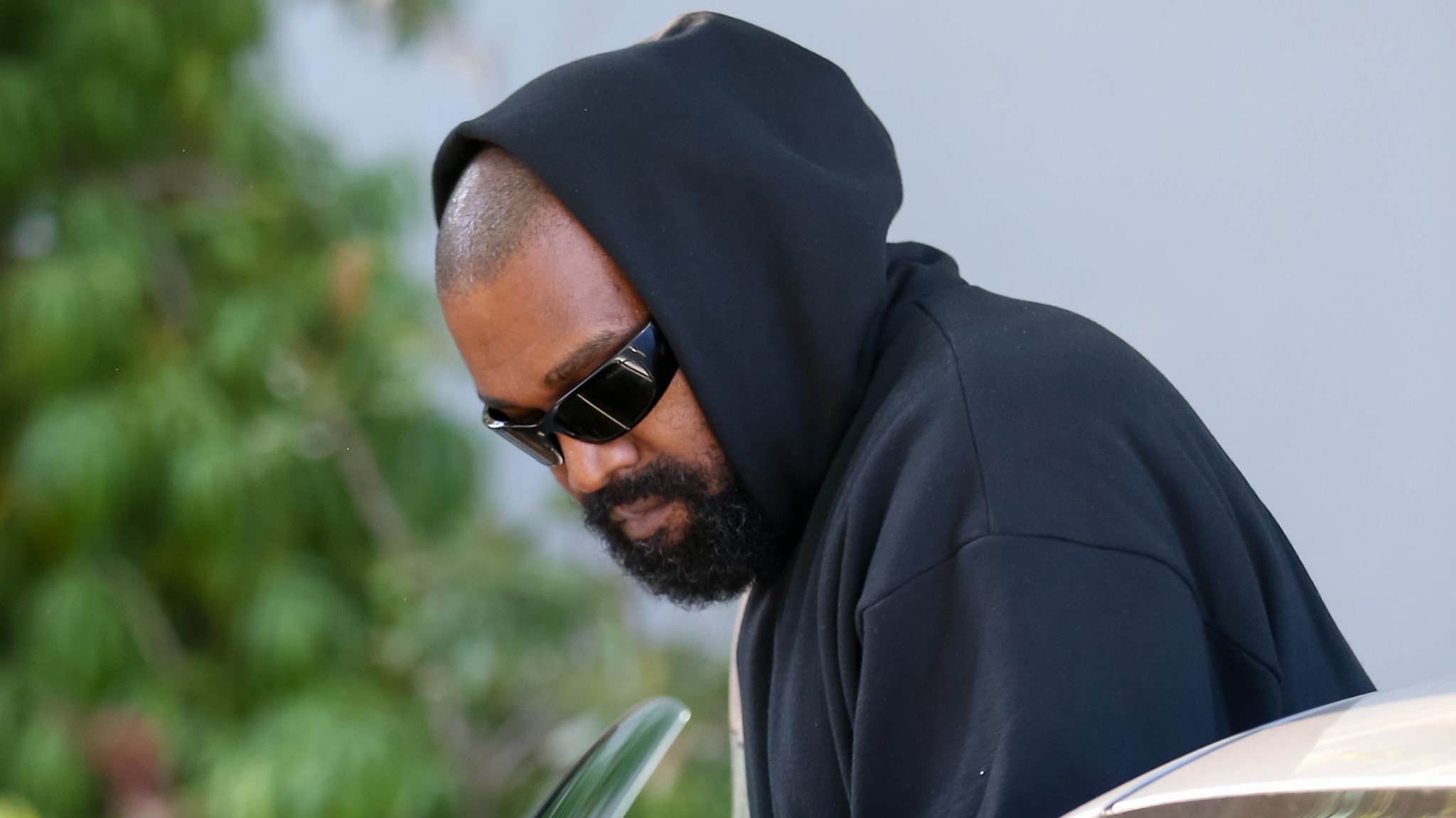 Ye wearing a black hoodie and sunglasses, looking down, with a blurred green background.