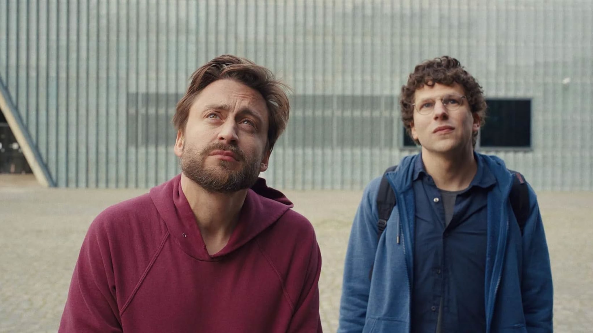 Kieran Culkin and Jesse Eisenberg stand outside, looking upwards. The man on the left wears a red hoodie, and the man on the right wears a blue jacket.
