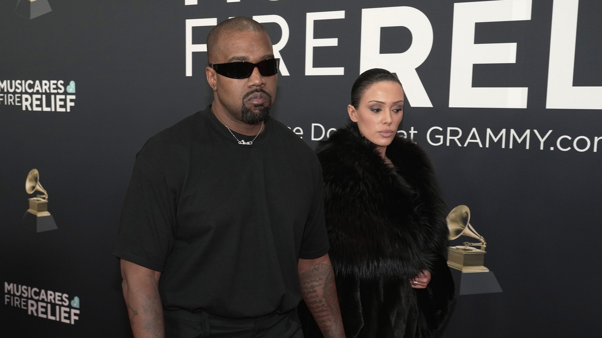 Kanye West in sunglasses and black attire, accompanied by a Bianca Censori in a fur coat, at a MusiCares event backdrop.