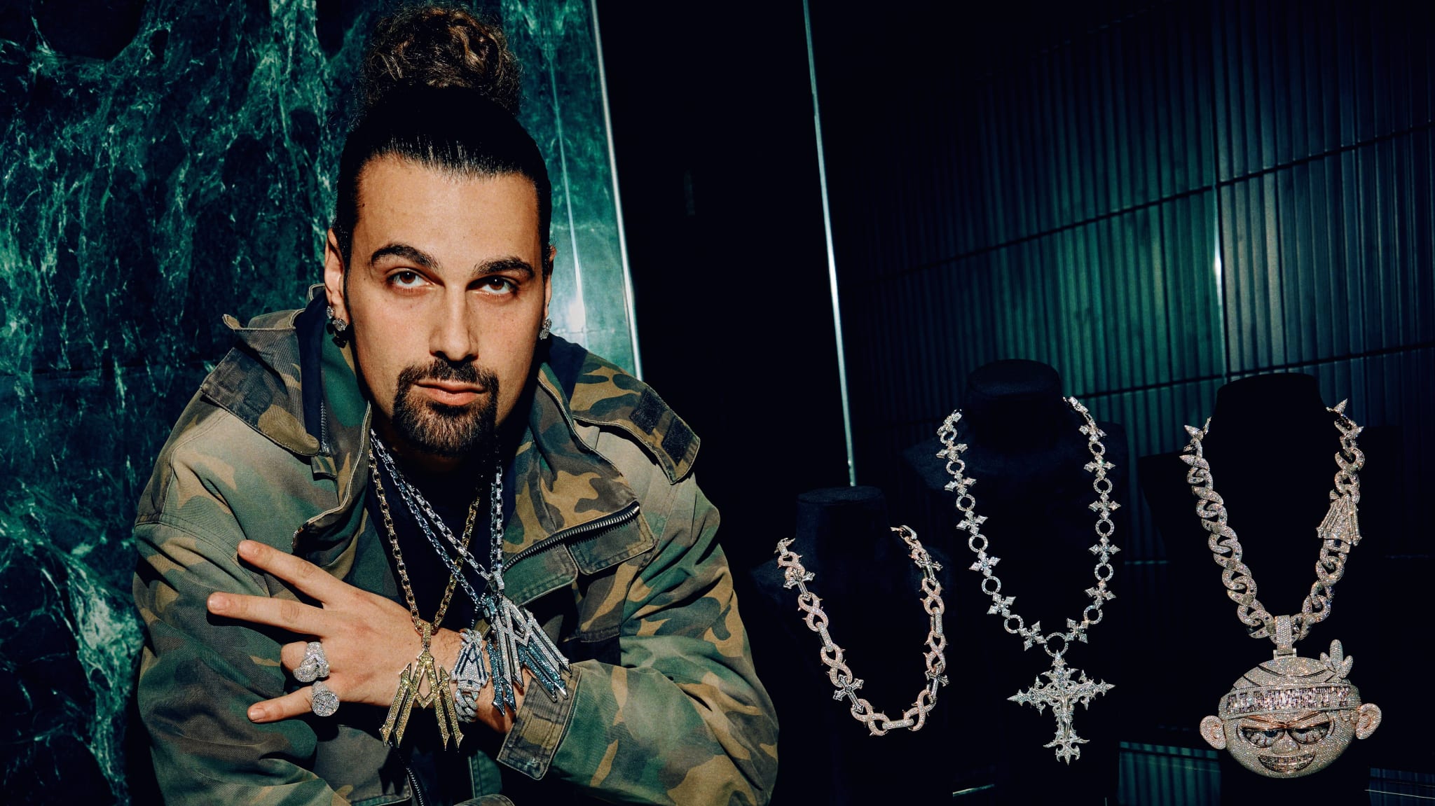 Alex Moss wearing a camouflage jacket, poses beside displays of ornate silver necklaces in a dimly lit room.