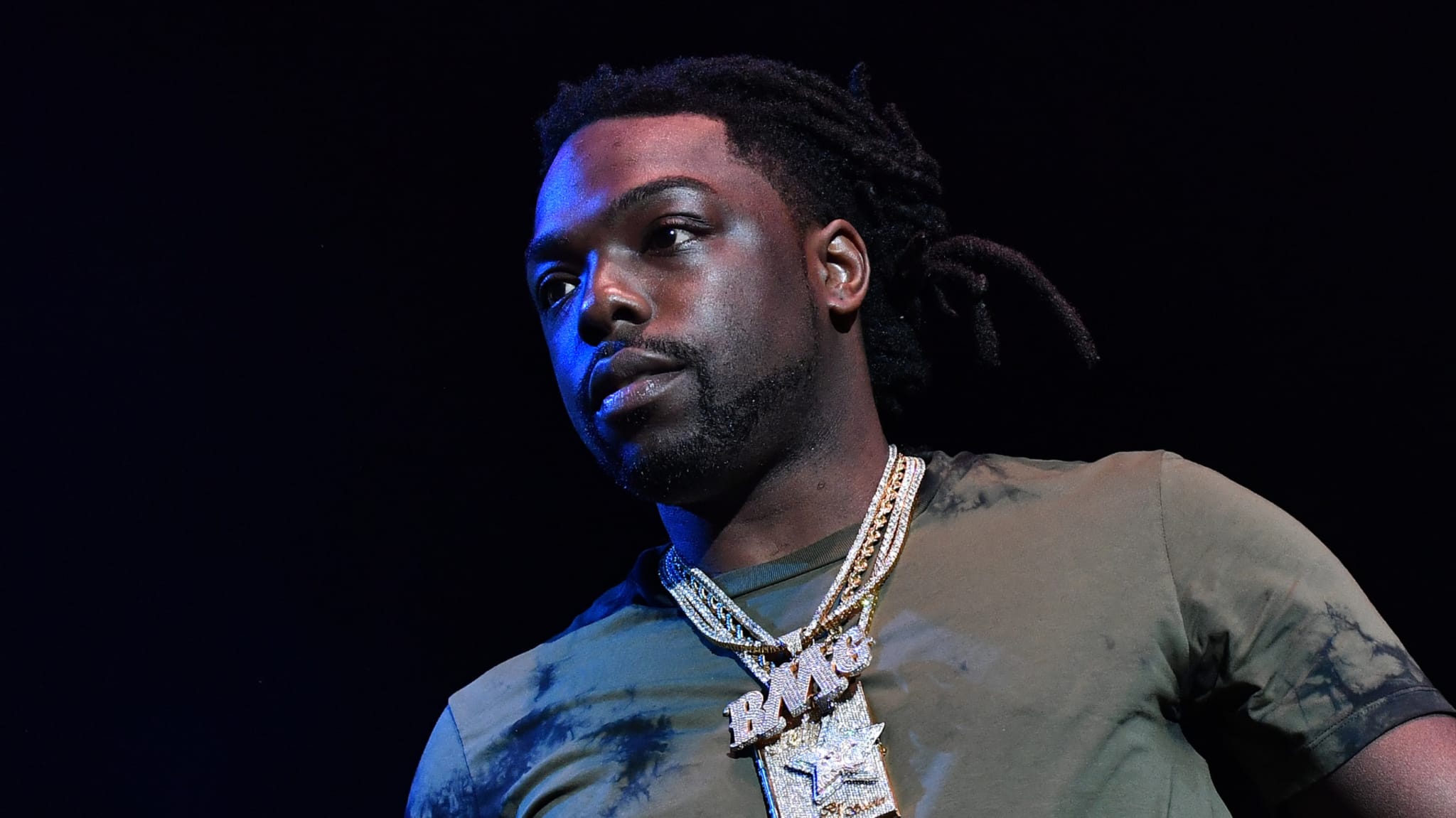 A man with dreadlocks and a beard, wearing a green shirt and multiple gold chains, stands against a dark background.