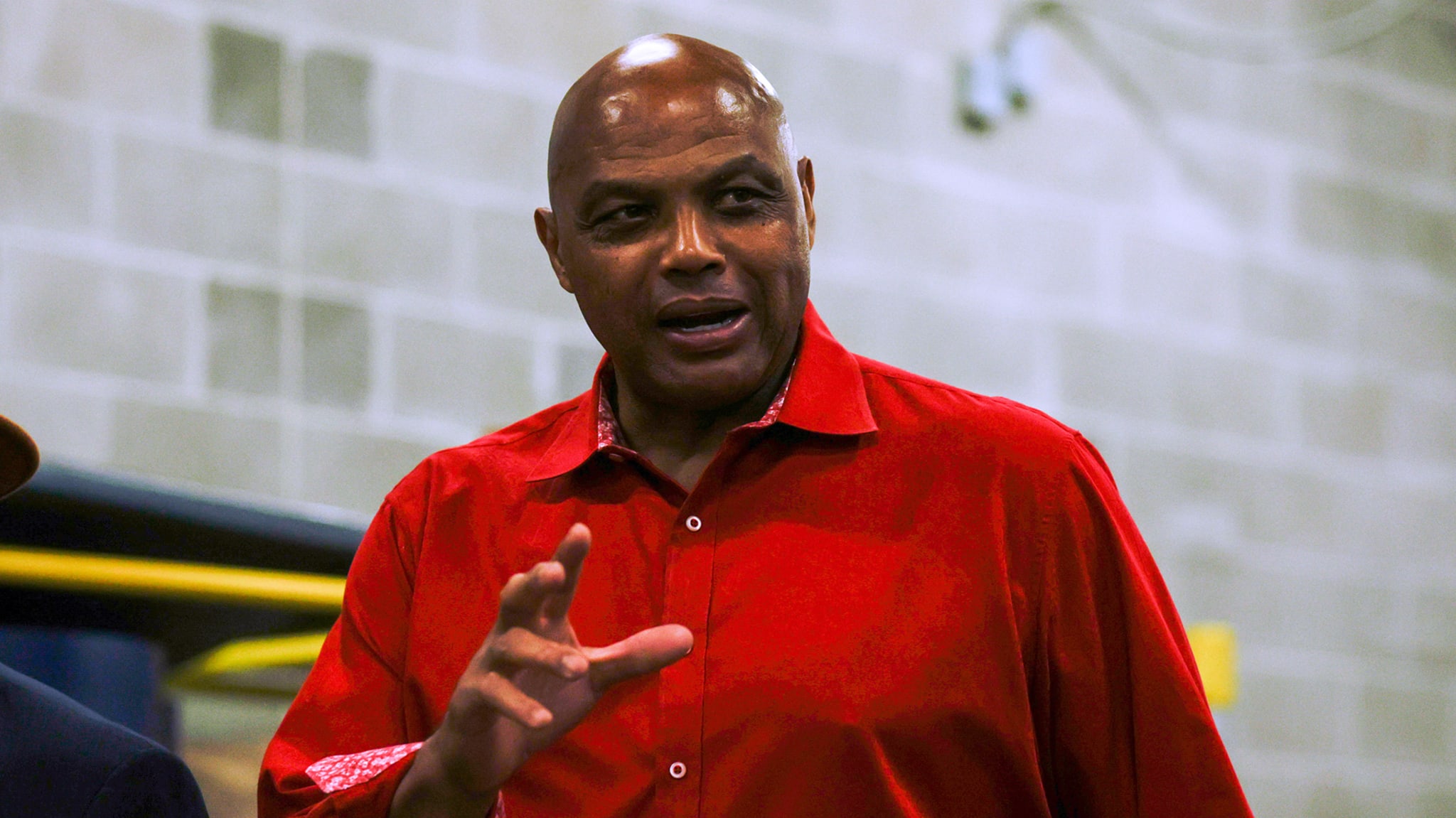Charles Barkley in a red shirt, gesturing while speaking, with a brick wall background.