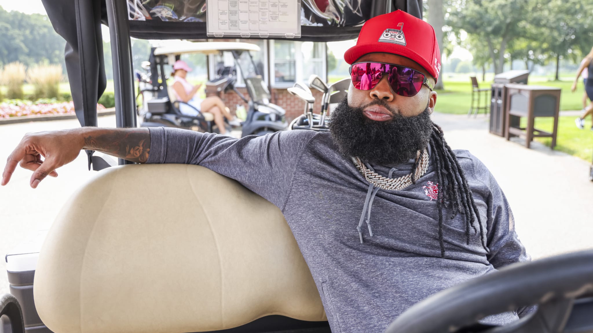 Sada Baby with a long beard and sunglasses sits in a golf cart, wearing a red cap and casual attire.