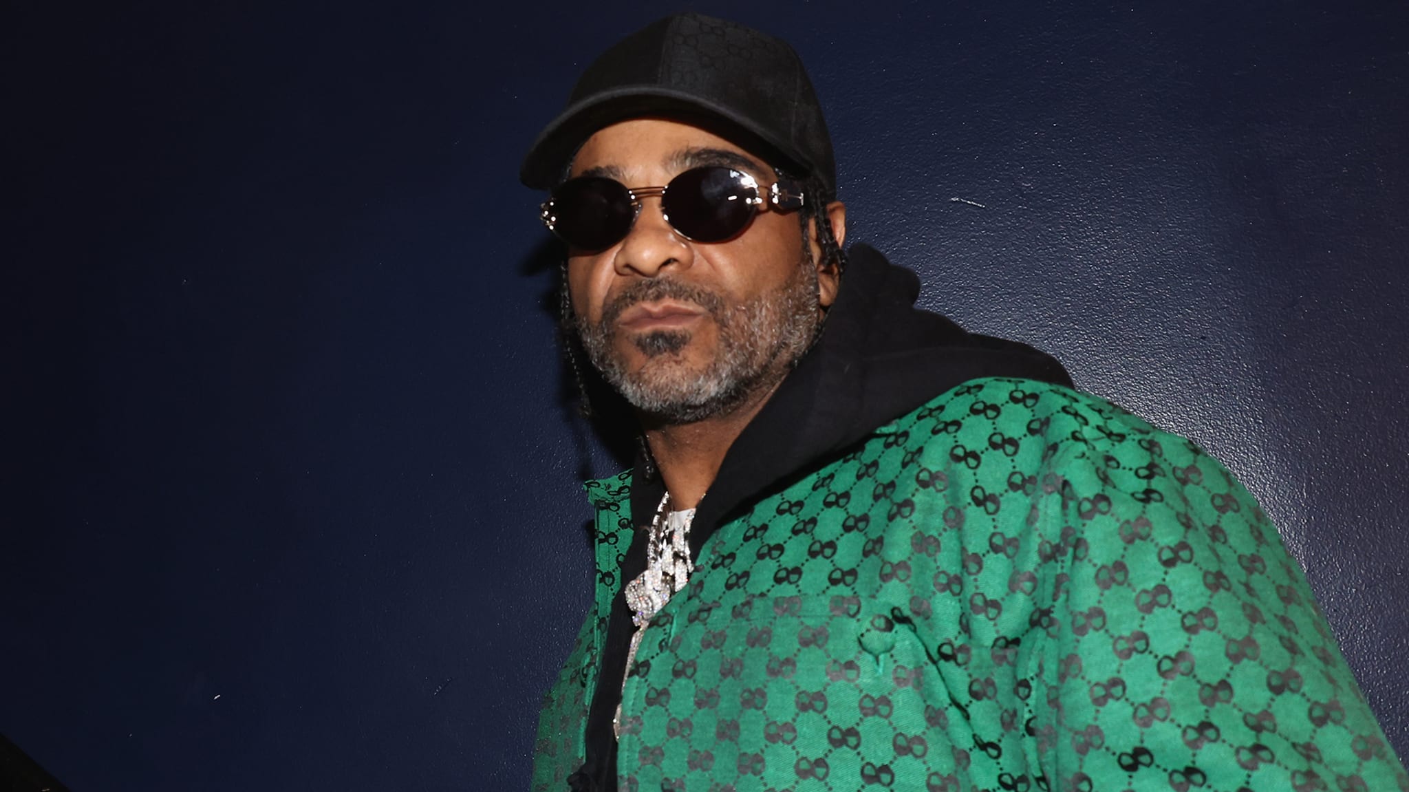 Jim Jones in a green Gucci jacket, sunglasses, and a black cap, standing against a dark blue background.