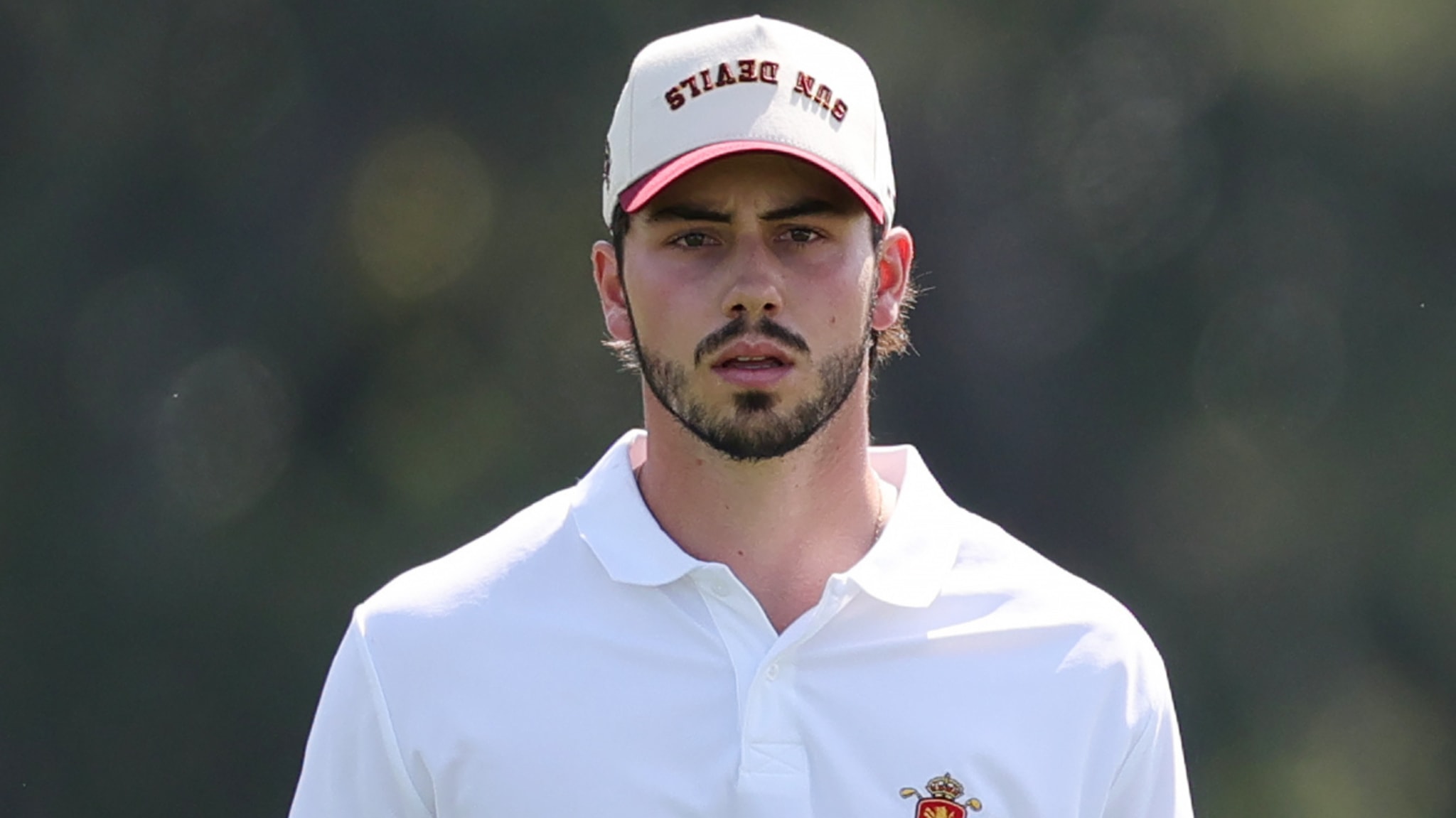 A man wearing a white polo shirt and a cap with text, standing outdoors.