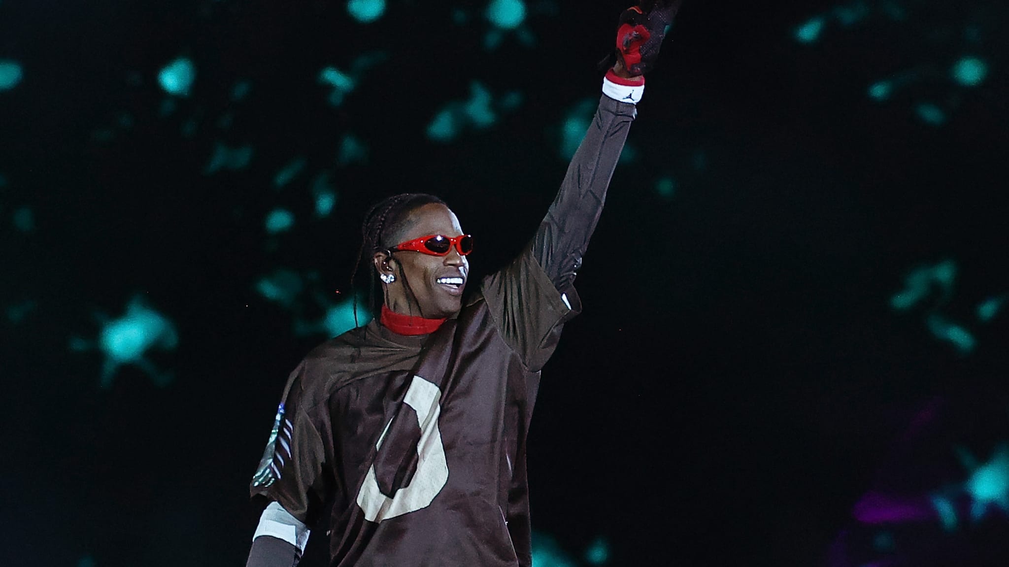 Travis Scott on stage wearing a dark outfit and red gloves, with braided hair and sunglasses, raising one arm against a starry backdrop.