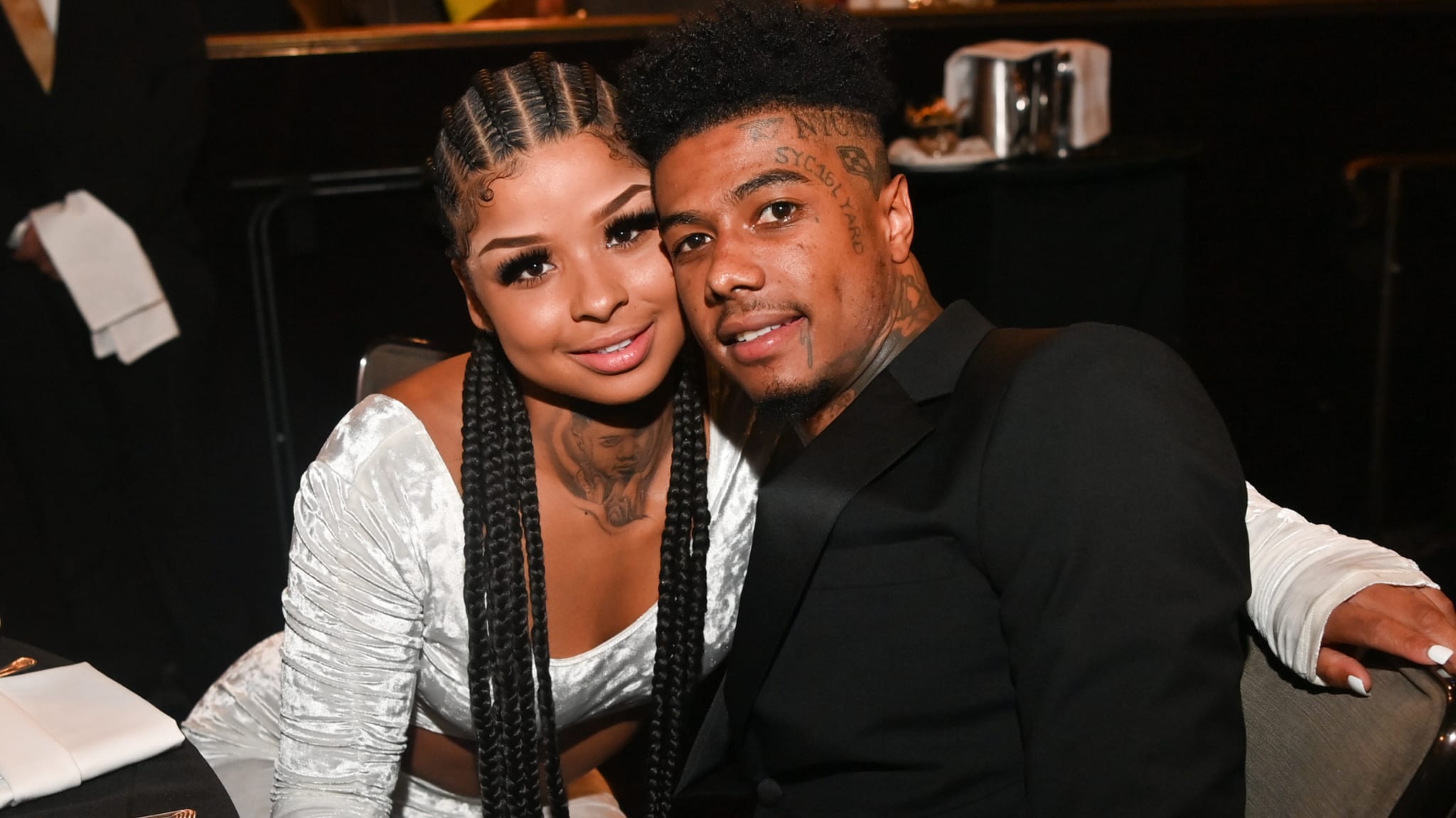 A couple posing together at an event. The woman has braided hair, and the man has tattoos on his face. They are sitting closely.