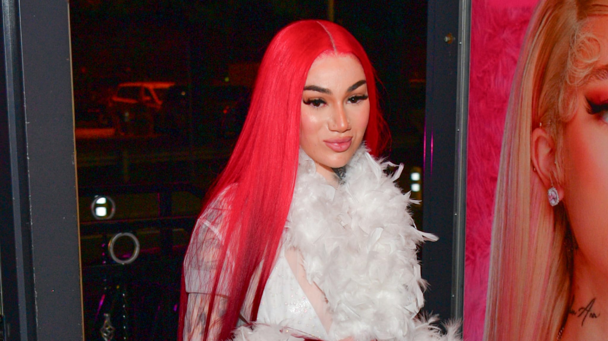 Bhad Bhabie with long red hair and a white feather boa stands in front of a large poster. The background is dimly lit.
