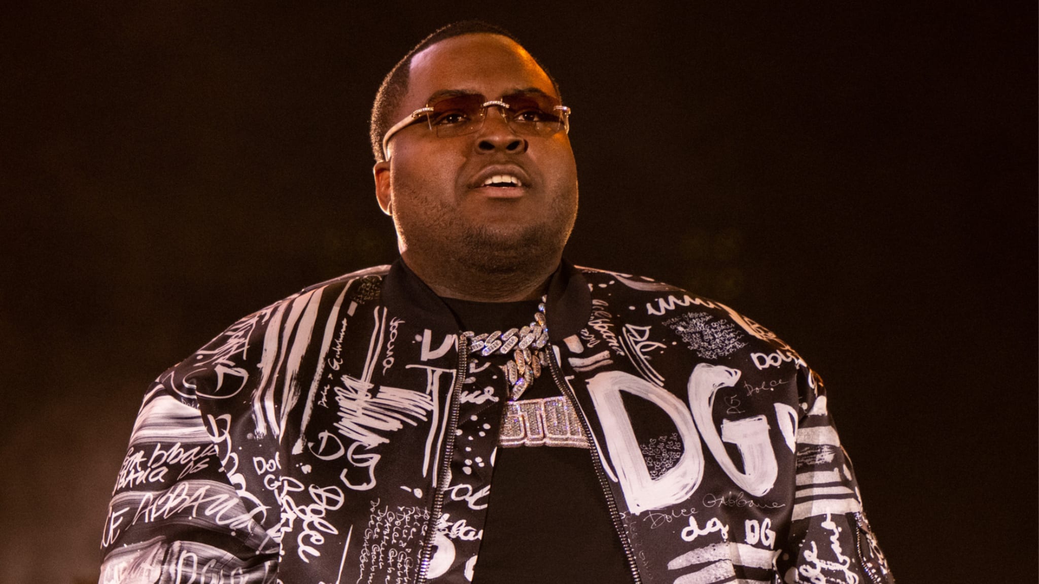 A man wearing sunglasses and a patterned jacket performs on stage.