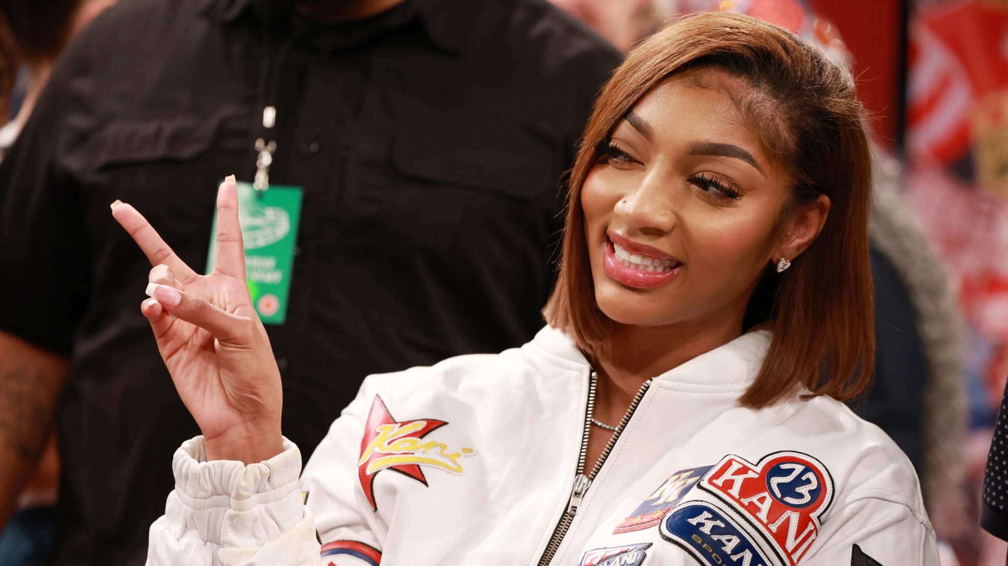 Angel Reese in a white jacket with patches, smiling and making a peace sign.