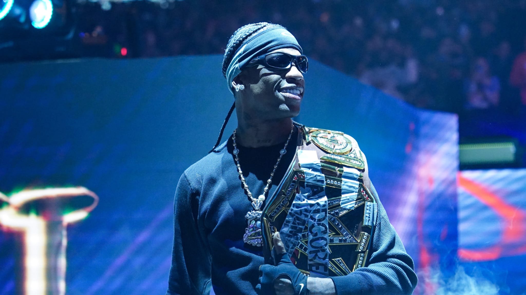 A person wearing sunglasses and a headband, holding a championship belt, stands confidently in a brightly lit arena.