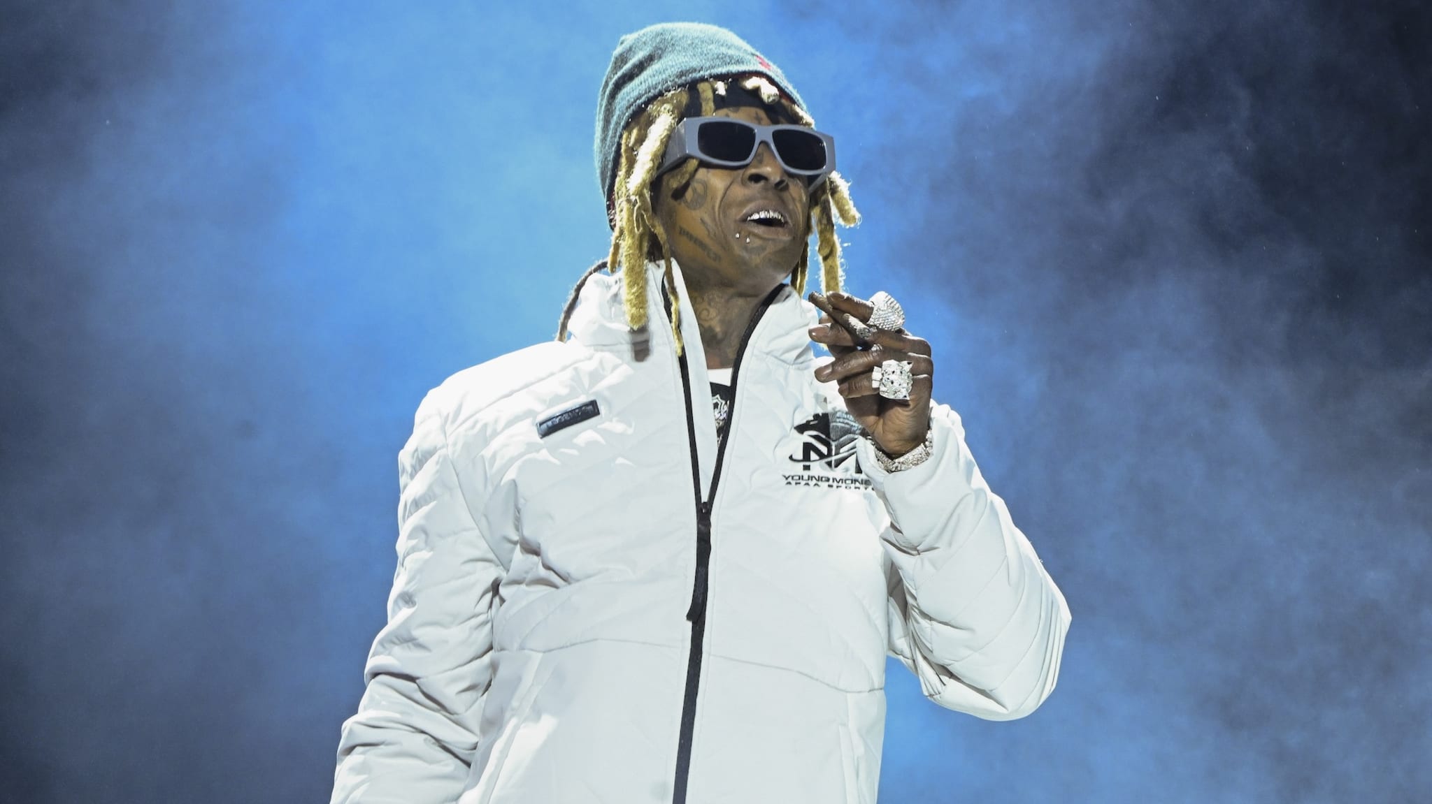 Lil Wayne performing on stage in a white jacket, sunglasses, and a beanie, with a microphone in hand against a smoky blue background.