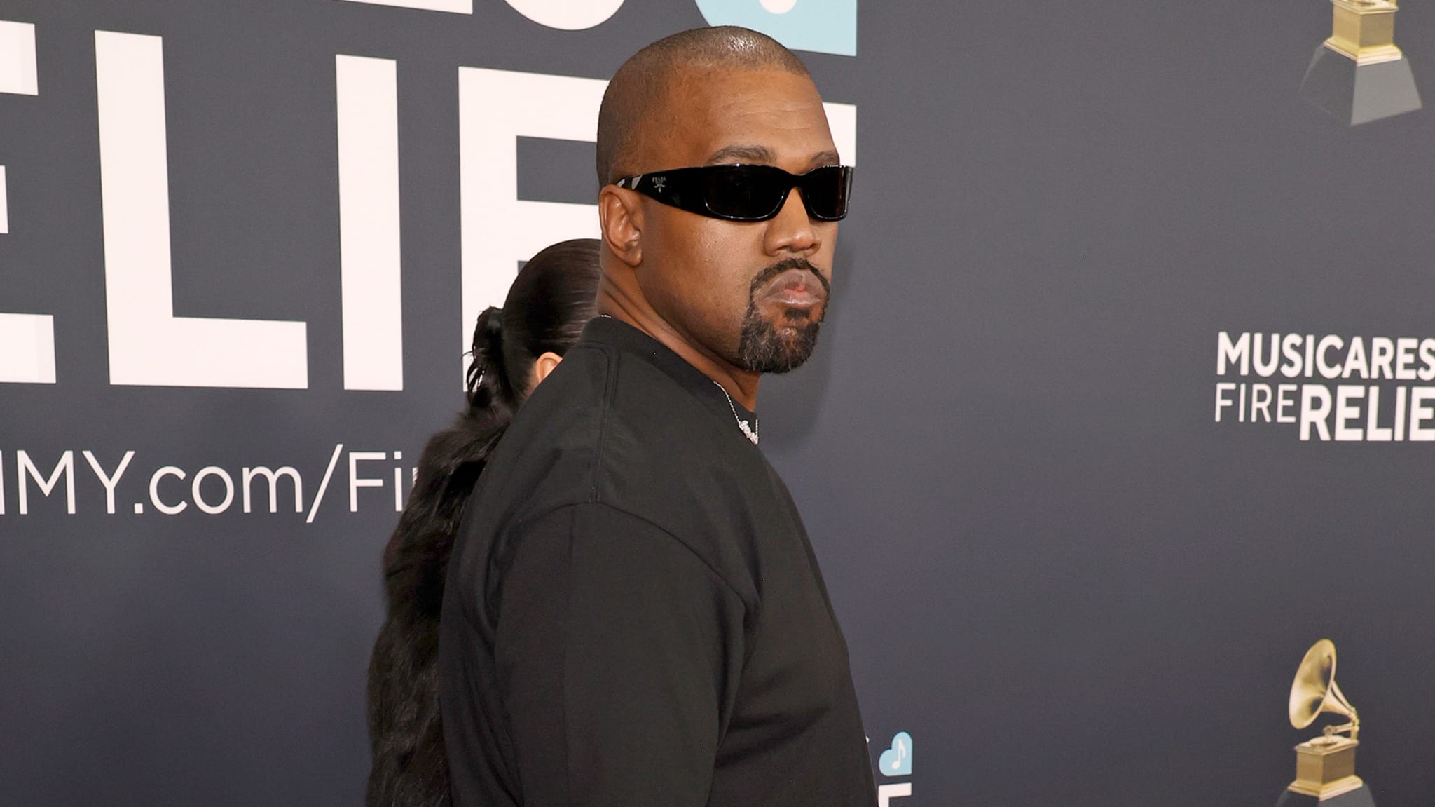 Kanye West wearing sunglasses and a black outfit at an event, standing in front of a backdrop with text and logos.