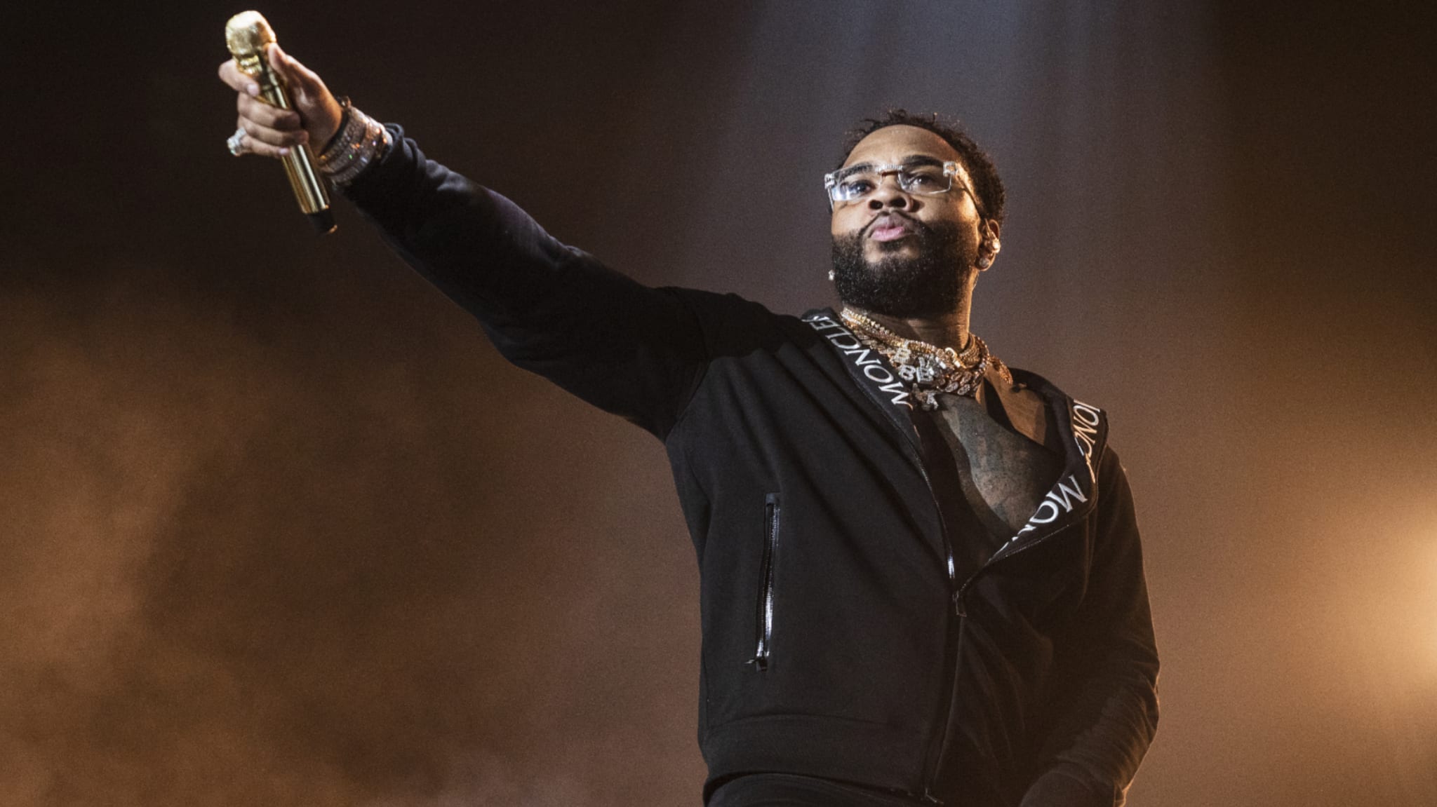 Kevin Gates on stage holding a microphone, wearing glasses and a black jacket, with dramatic lighting in the background.