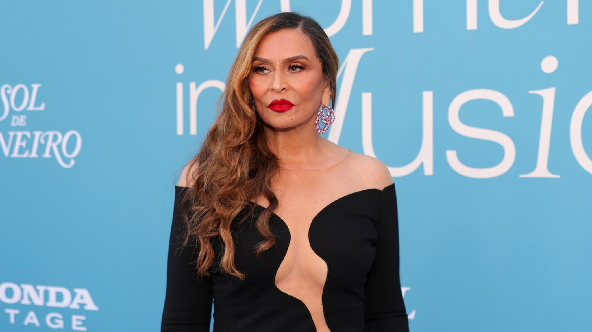 A woman with long hair in a black dress stands against a blue background with text, wearing red lipstick and earrings.