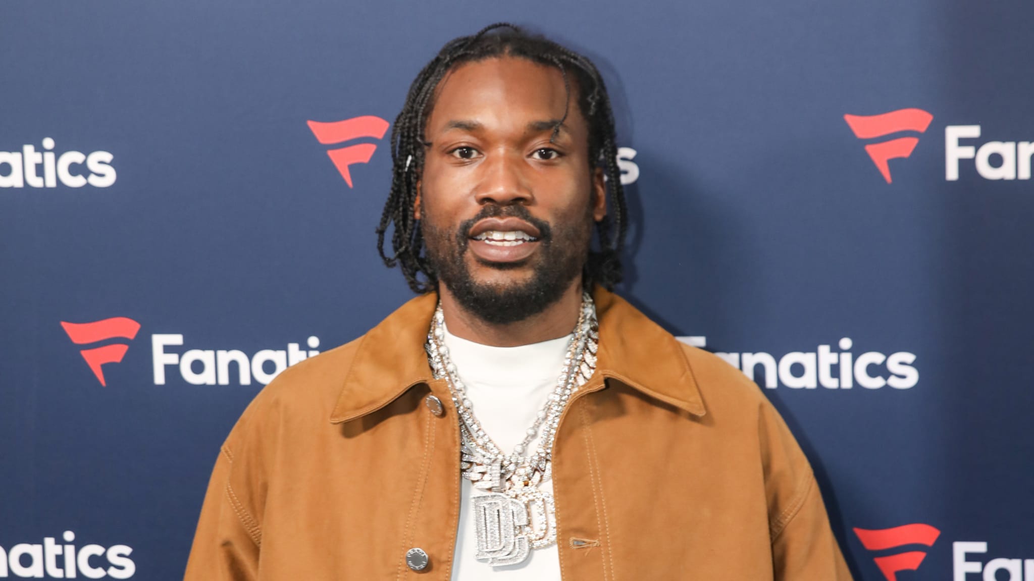 Meek Mill at a Fanatics event, wearing a brown jacket and a large chain necklace, smiling at the camera.