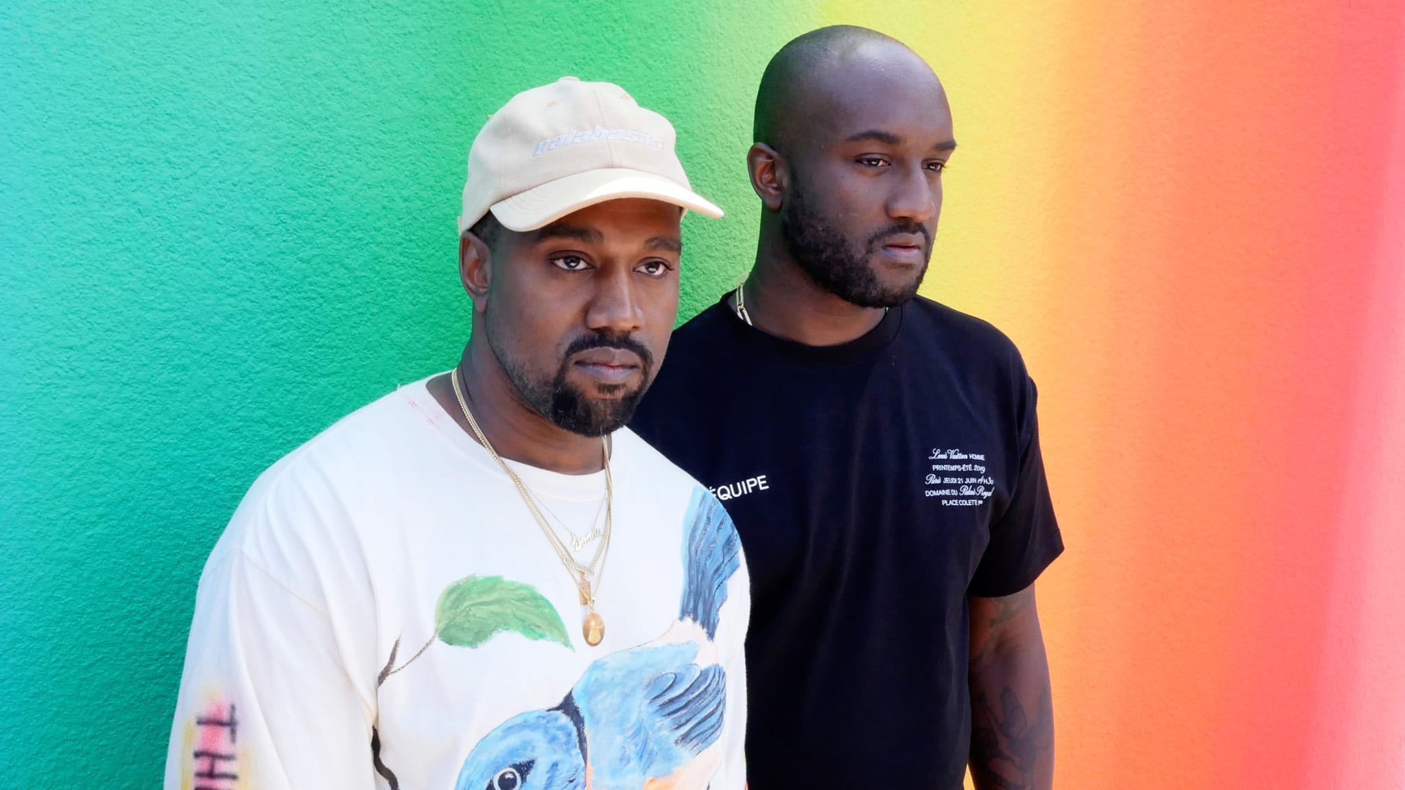 Kanye West and Virgil Abloh stand in front of a colorful gradient wall. Kanye wears a bird-print shirt and cap, Virgil in black tee.