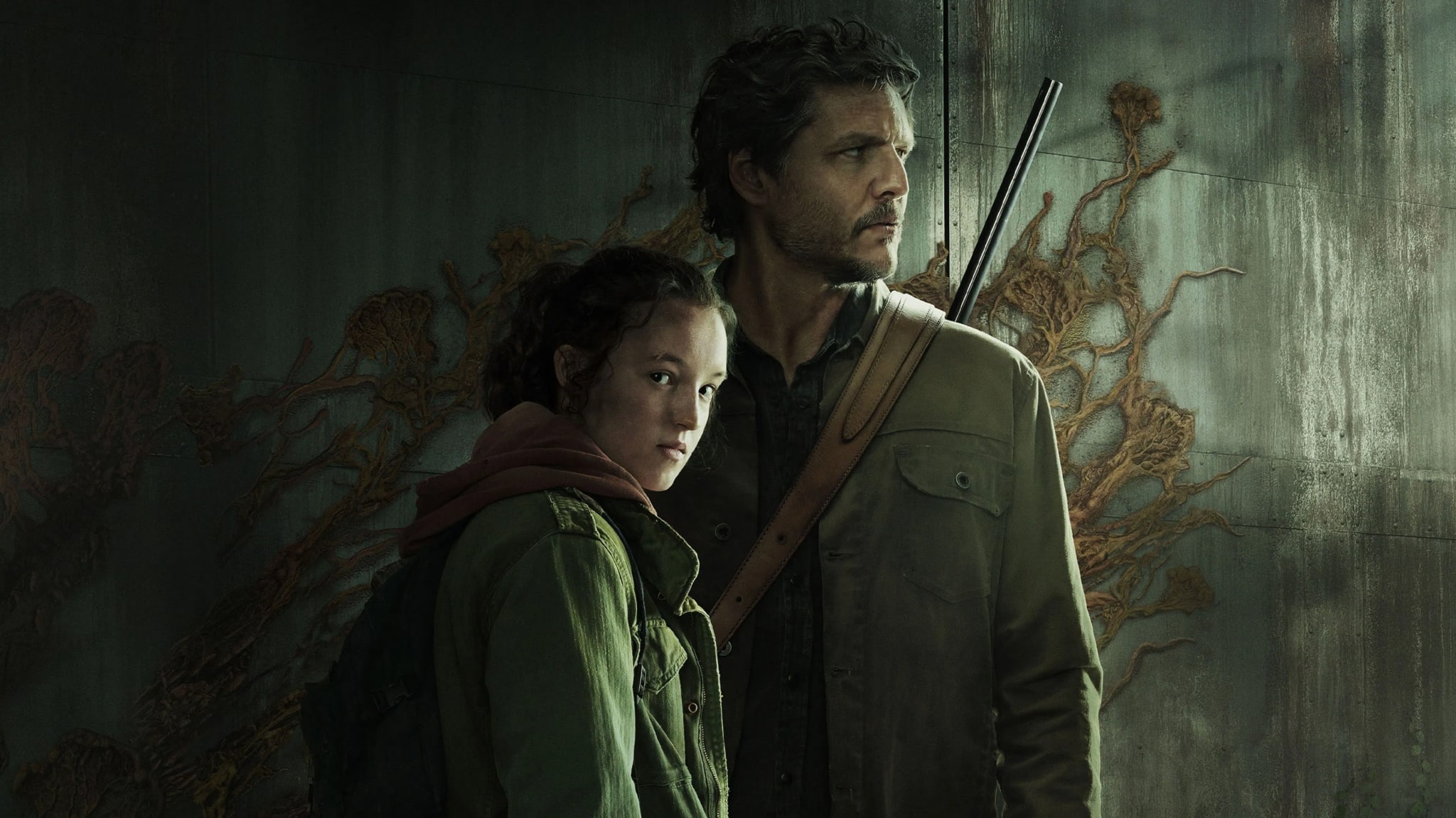 Pedro Pascal and Bella Ramsey in a scene from "The Last of Us," standing against a wall with fungal growths, looking serious.