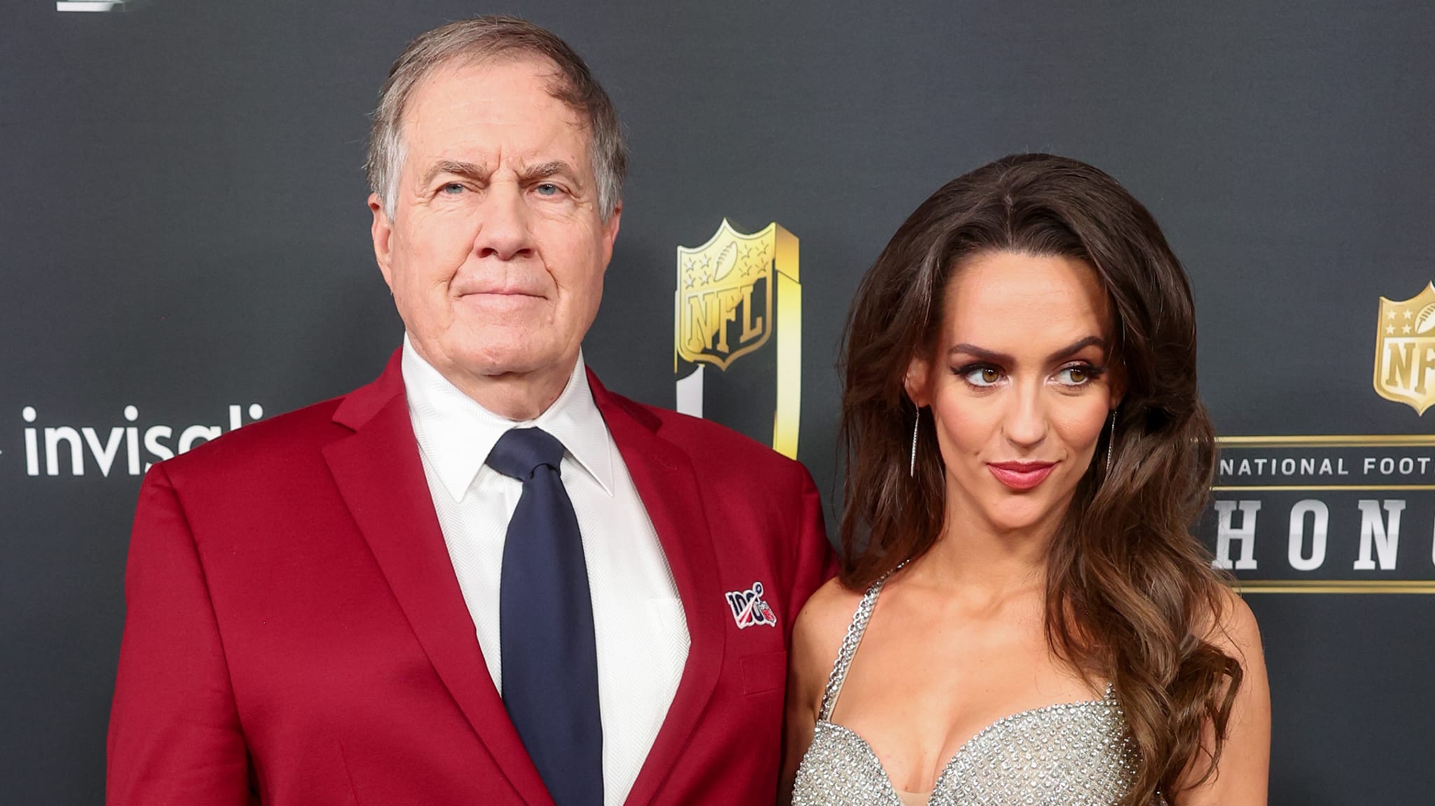Bill Belichick in a red suit and tie stands beside girlfriend Jordon Huston in a silver dress at the 14th Annual NFL Honors