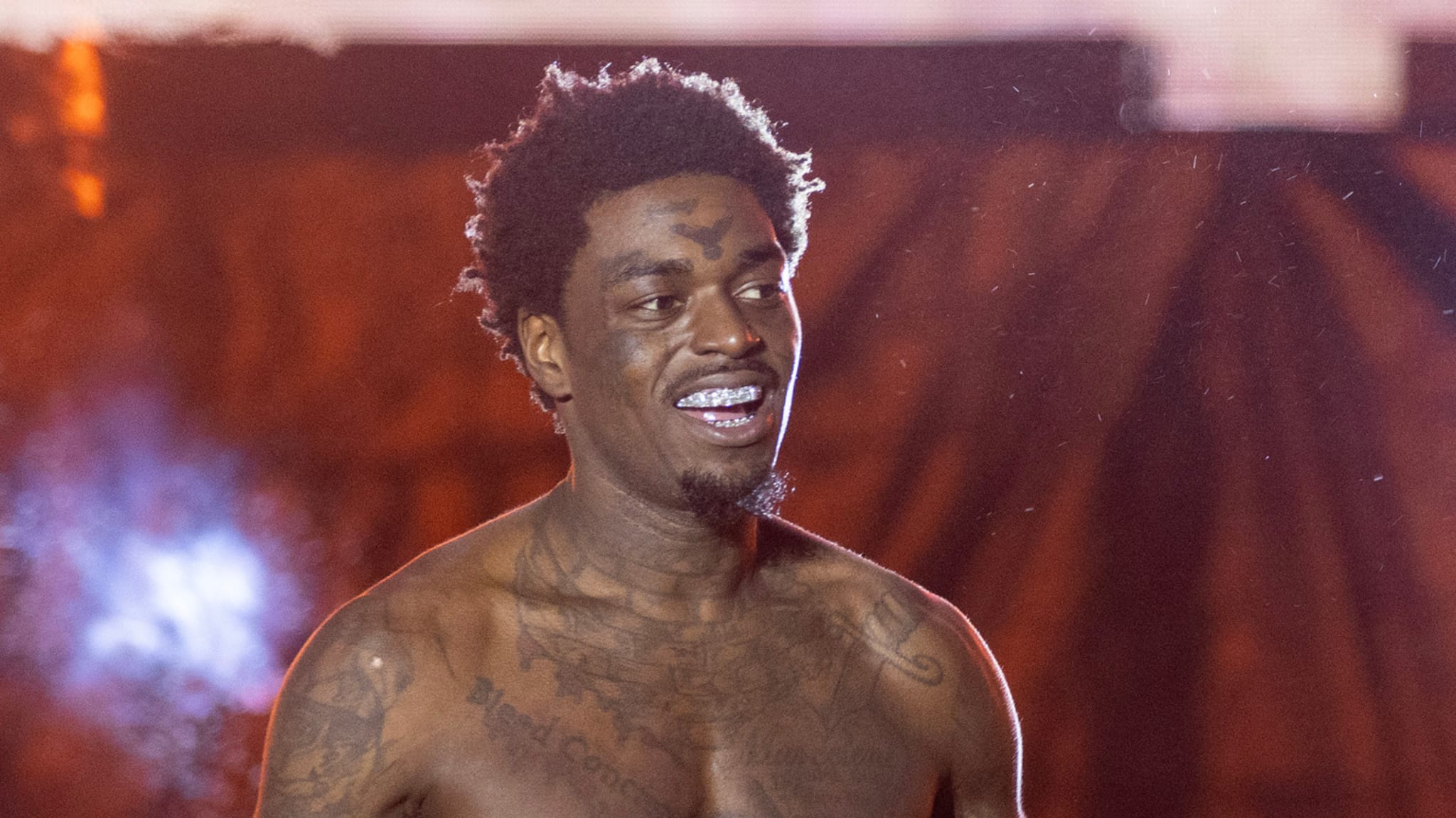 A shirtless man with tattoos and a grill smiles on stage with a blurred background.