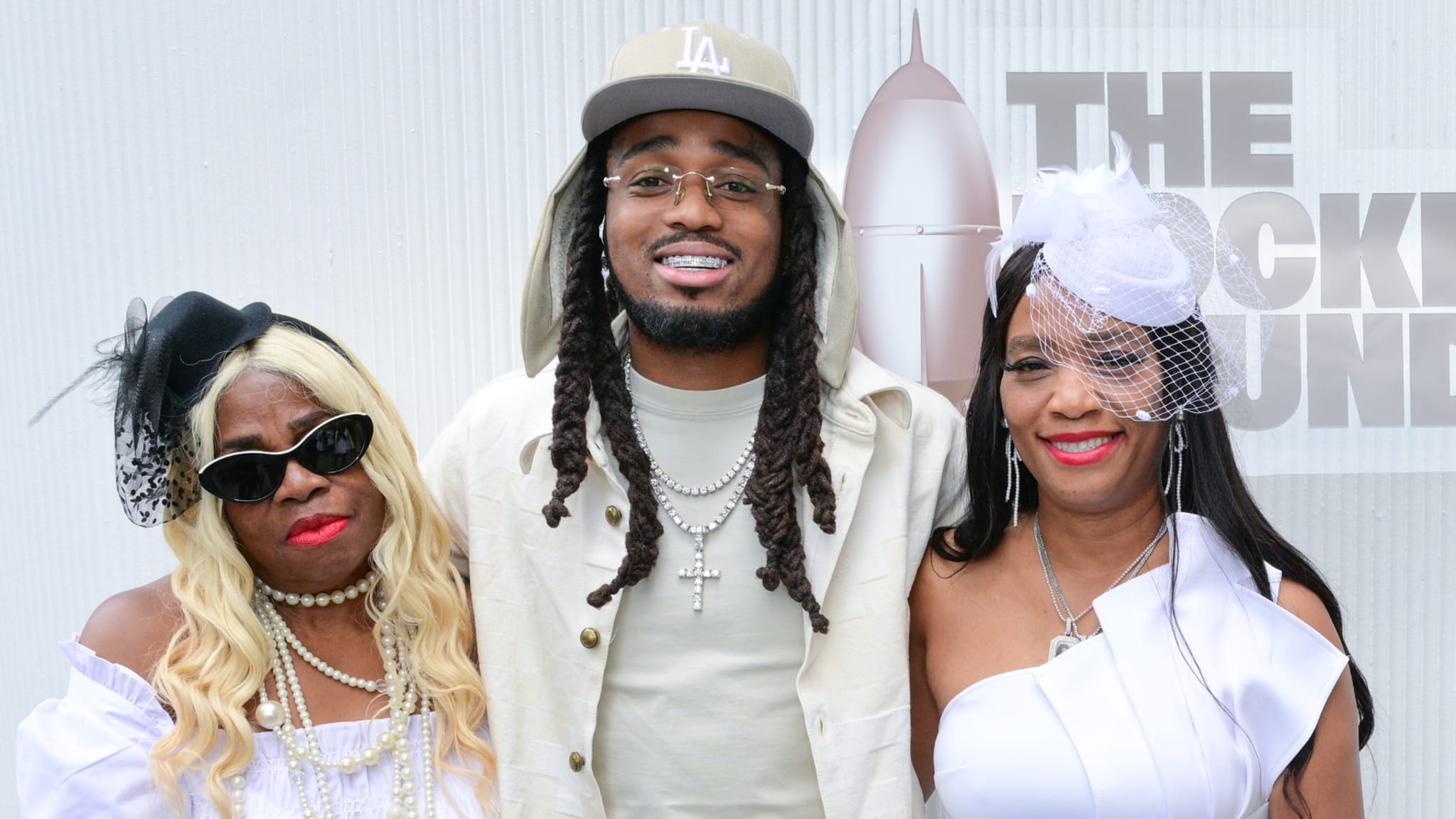 Quavo, wearing a beige hat and white outfit, stands between two women in white dresses and hats, smiling at an event.