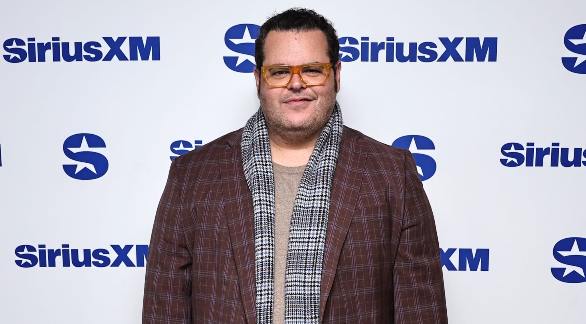 Josh Gad at a SiriusXM event, wearing a plaid jacket, scarf, and glasses, smiling at the camera.