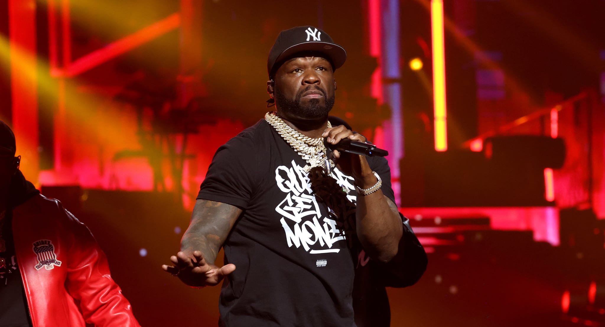 Rapper on stage performing, wearing a graphic tee, chain necklace, and cap