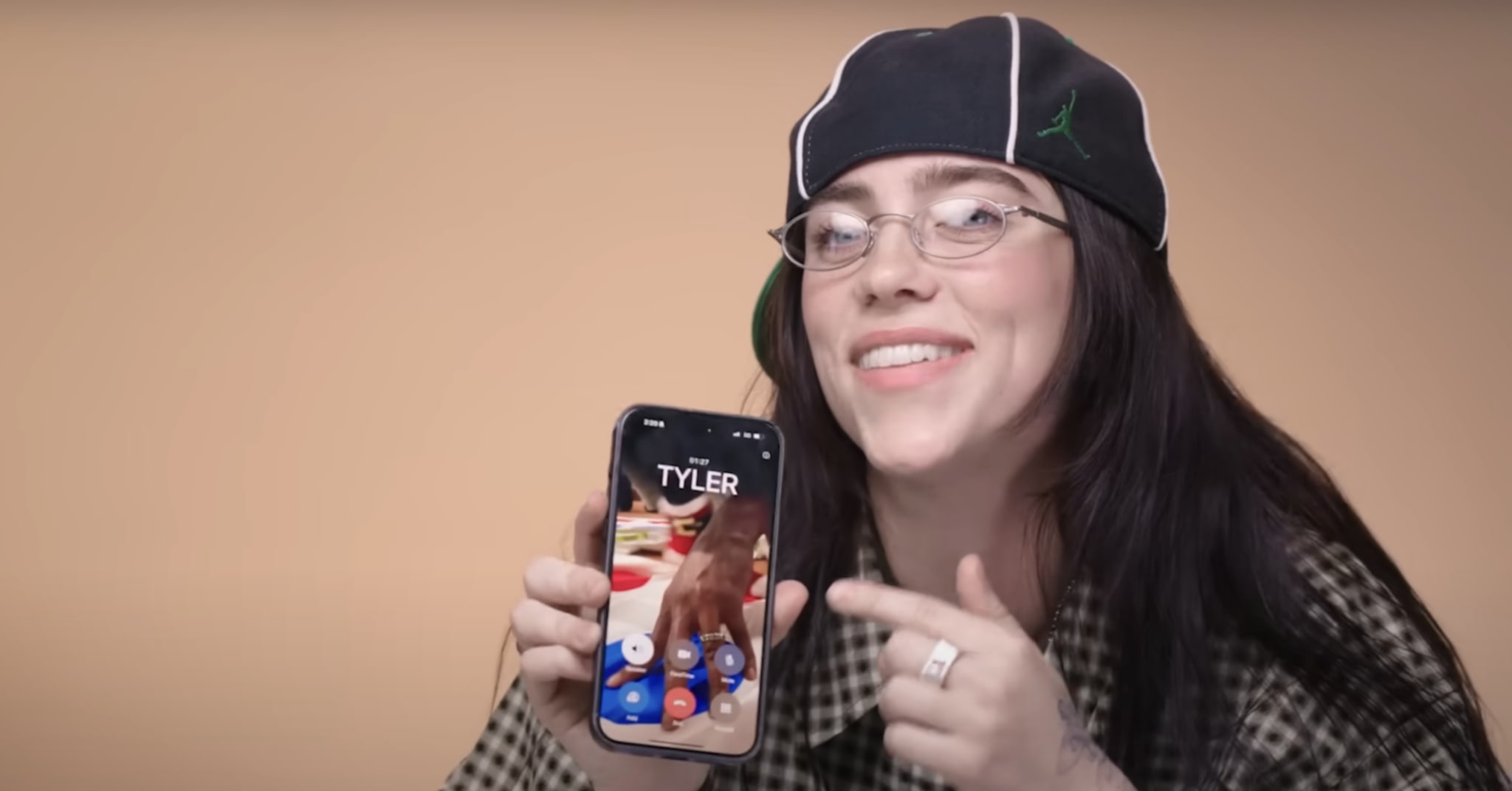 Billie Eilish, wearing a casual plaid jacket and a hat, smiles and shows her phone screen with an incoming call from Tyler