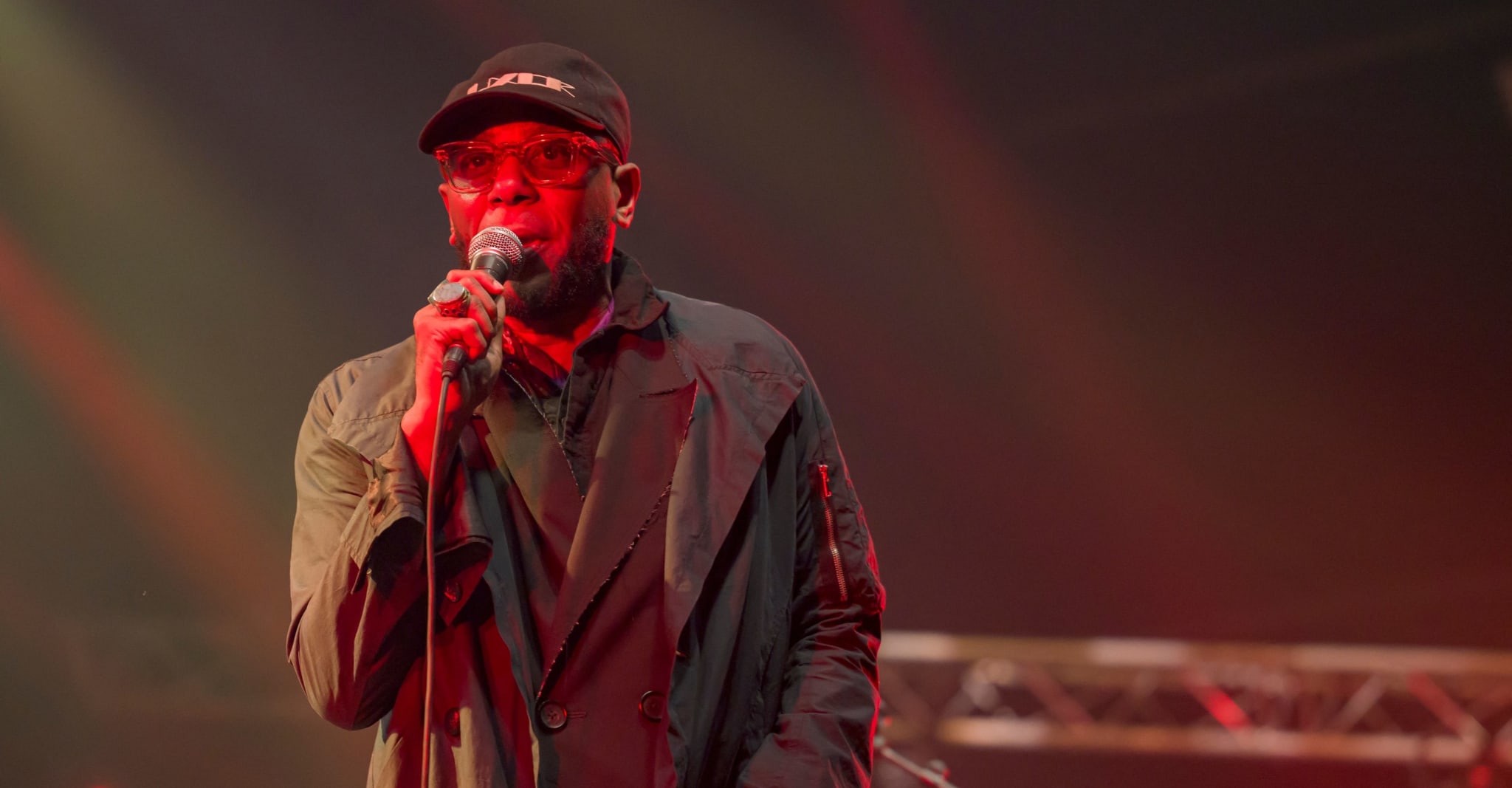 A man in a hat and dark, layered clothing performs on stage, holding a microphone