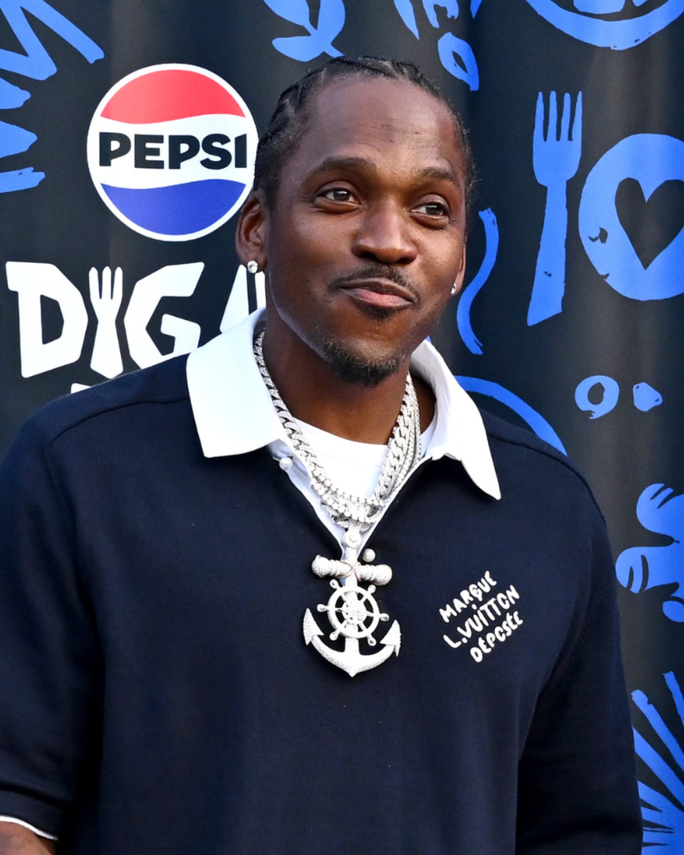 Two men stand in front of a Pepsi backdrop, both smiling. They wear black shirts and large chains.