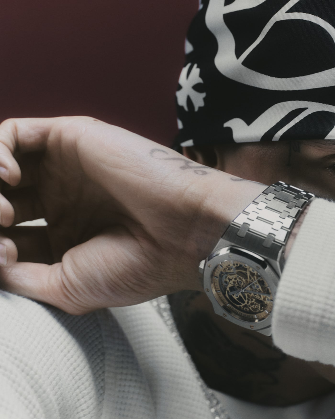 A person wearing a patterned bandana and a silver watch, partially covering their face with their arm, dressed in a white sweater.