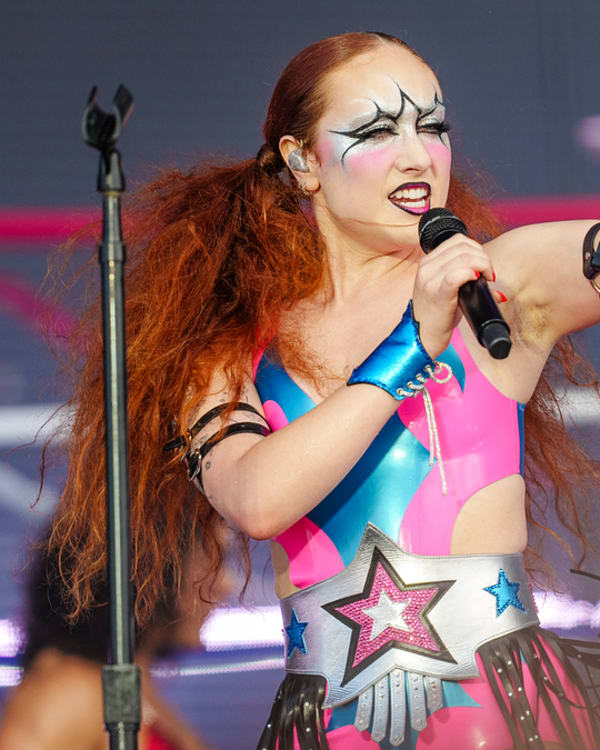 Chappell Roan  wearing a luchador costume with star designs, performs passionately on stage, holding a microphone. Gymnasts can be seen in the background