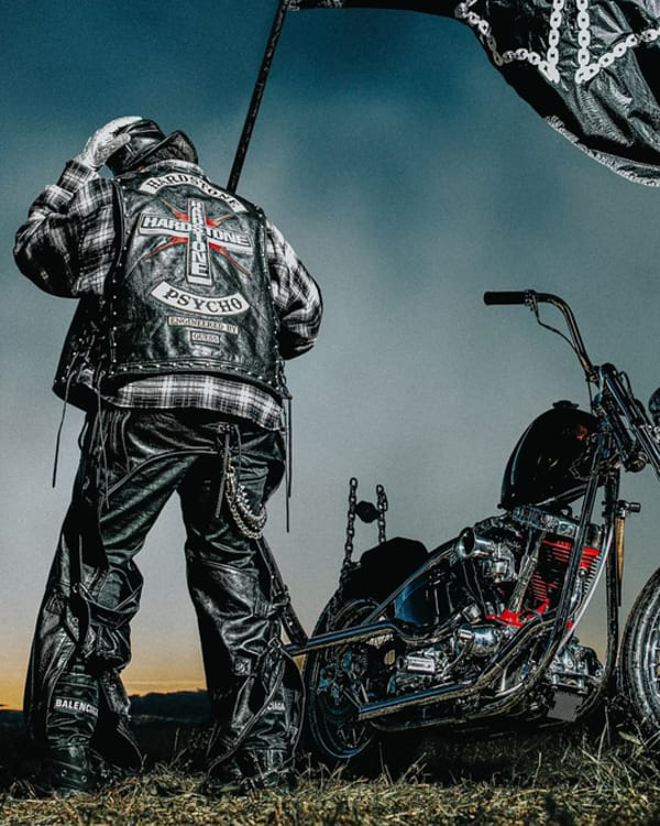 A biker in leather gear stands by a custom motorcycle, holding a flag. The back of the biker's jacket reads "Hardcore Carnivore Psycho." The image is related to music