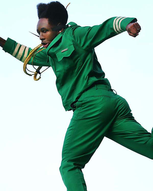 Tyler in a green suit mid-jump, holding a microphone, with an energetic pose against a plain background.