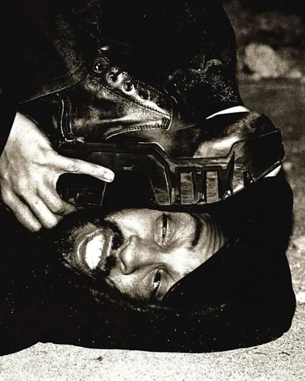 A man lying on the ground with a boot pressed against his face, smiling. The image is in black and white.