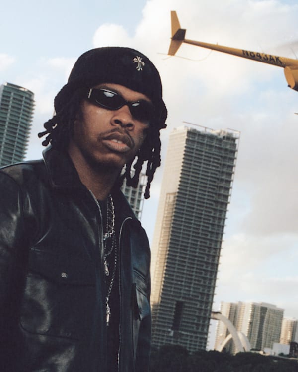 A man in a black leather jacket and sunglasses stands in front of city skyscrapers. A yellow helicopter flies overhead.