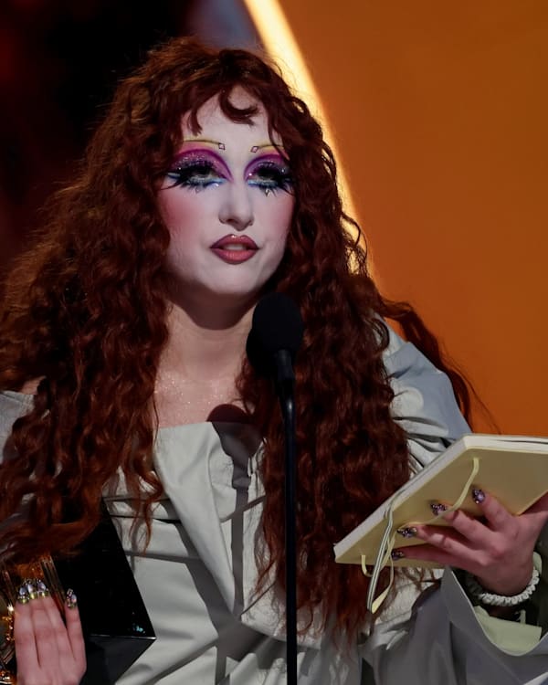 A person with long red hair and dramatic makeup holds a Grammy award and a notebook, speaking at a microphone on stage.