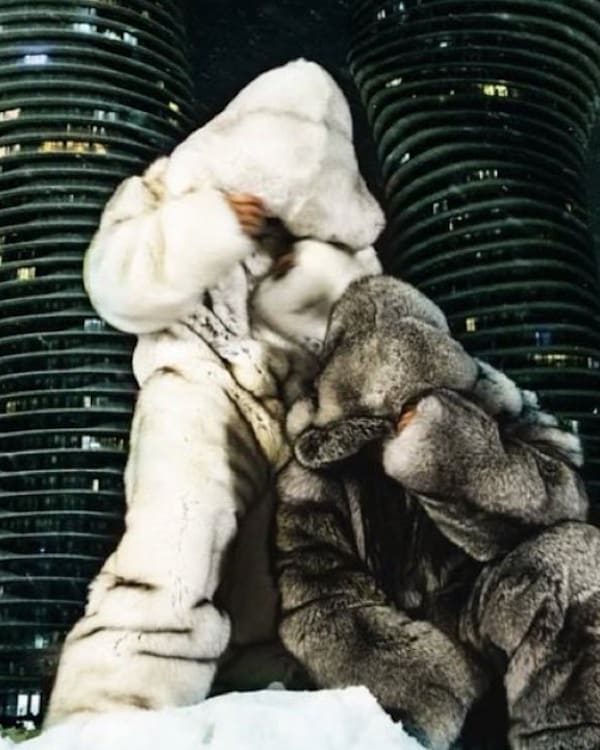 Two people in large fur outfits pose in front of curved skyscrapers at night, surrounded by snow.