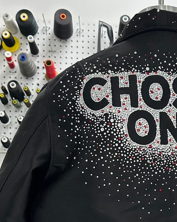 A black jacket with "CHOSEN ONE" in bold letters, adorned with a pattern of white and red dots. Sewing supplies are visible in the background.